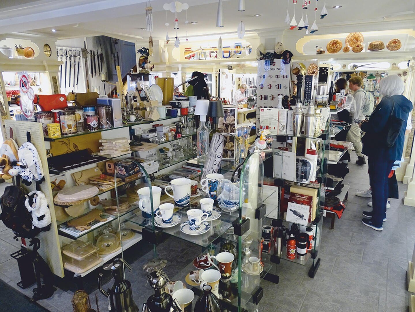 Ein Ladeninterieur mit Regalen voller Küchengeräte, Keramik und Dekorationsartikel. Zwei Personen stöbern in den Auslagen, die Tassen, Teekannen, Utensilien und andere Haushaltswaren umfassen.