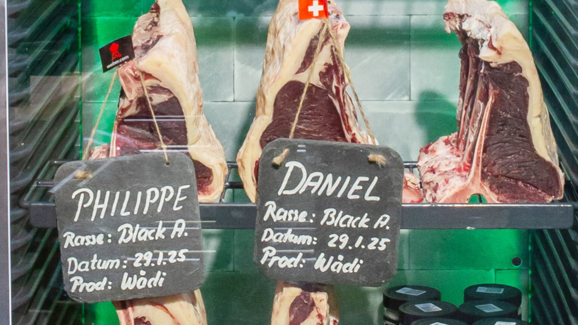 Trois grandes pièces de bœuf sont présentées verticalement dans une vitrine réfrigérée. Deux ardoises, intitulées PHILIPPE et DANIEL, indiquent la race, la date et les coordonnées du producteur en écriture blanche.