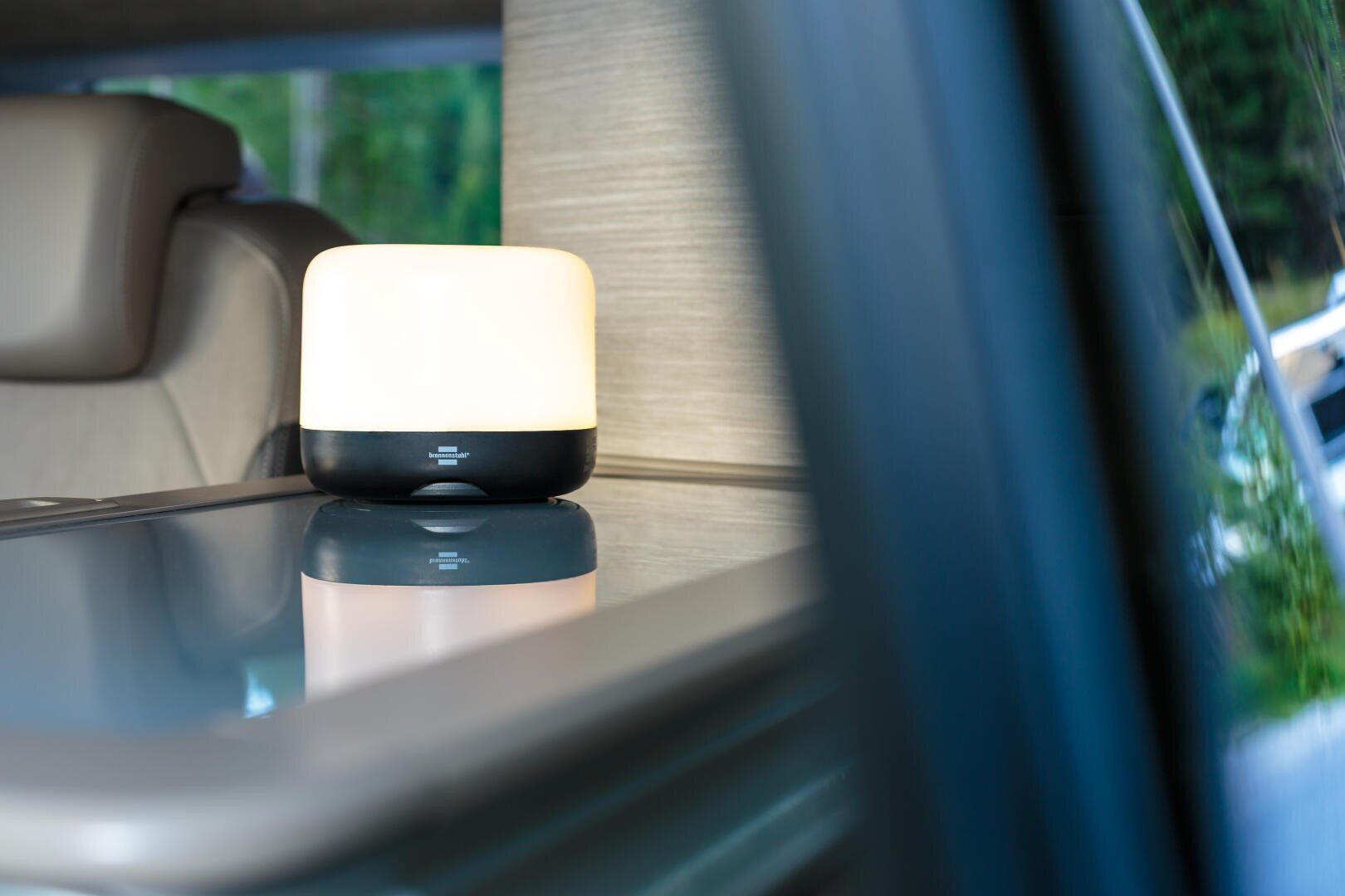 Eine kleine, moderne Lampe mit weißem Lampenschirm und schwarzem Sockel steht auf einer spiegelnden Oberfläche im Innenraum eines Fahrzeugs, in der Nähe eines grauen Sitzes und einer Holzverkleidung.