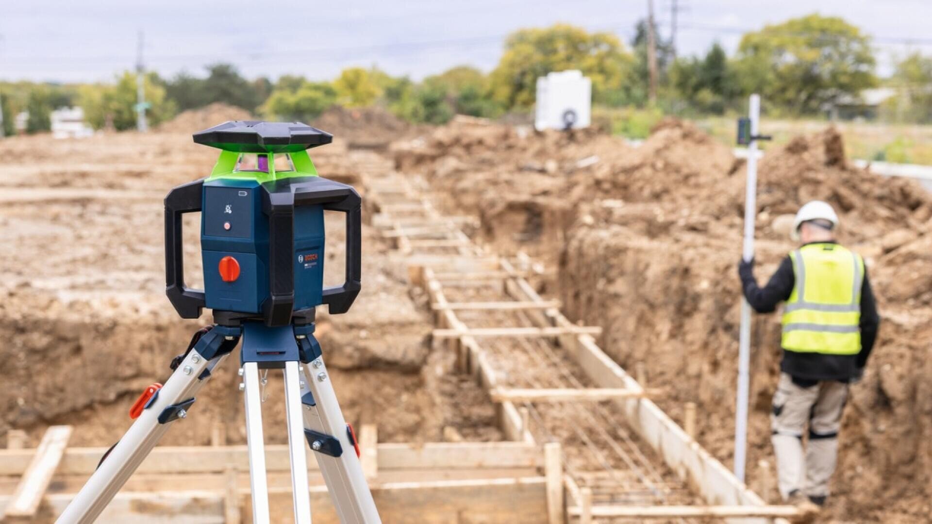 Im Vordergrund ein Bosch-Rotationslaser auf einem Stativ, im Hintergrund ein Bauarbeiter, der eine Messlatte in der Hand hält.