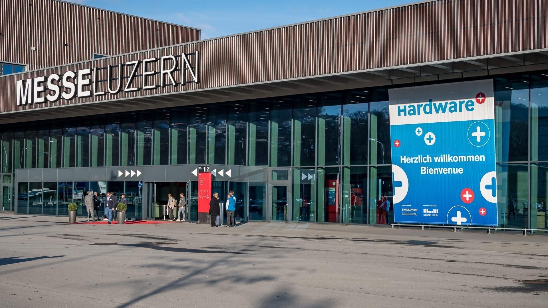 Vor dem Eingang der Messe Luzern, einem modernen Gebäude mit großen Glasfenstern, stehen Menschen. Auf einem blauen Schild steht „Hardware“ und „Herzlich willkommen – Bienvenue“. Die Szene ist hell und klar.
