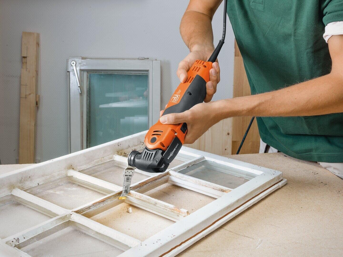 Eine Person bearbeitet mit einem orangefarbenen elektrischen Multifunktionswerkzeug den Holzrahmen eines alten Fensters und entfernt oder schneidet dabei einen Teil des Rahmens auf einer Werkbank ab.