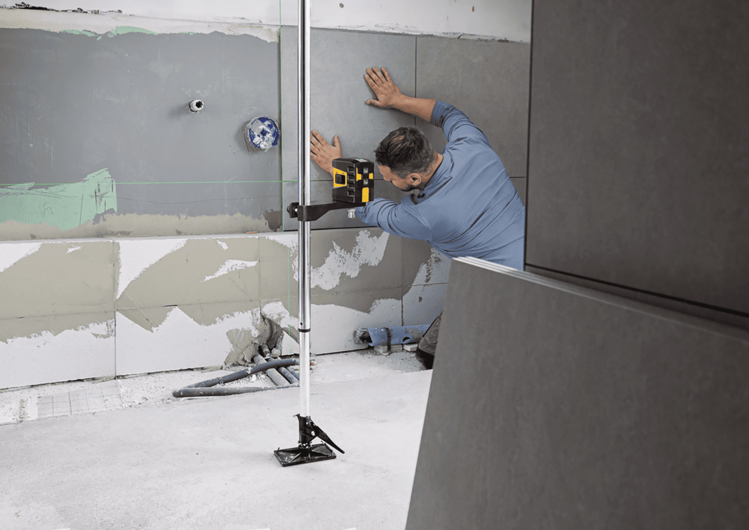 Ein Fliesenleger verwendet eine an einer Teleskopstange befestigte Laserwasserwaage, um eine graue Fliese an der Wand auszurichten.