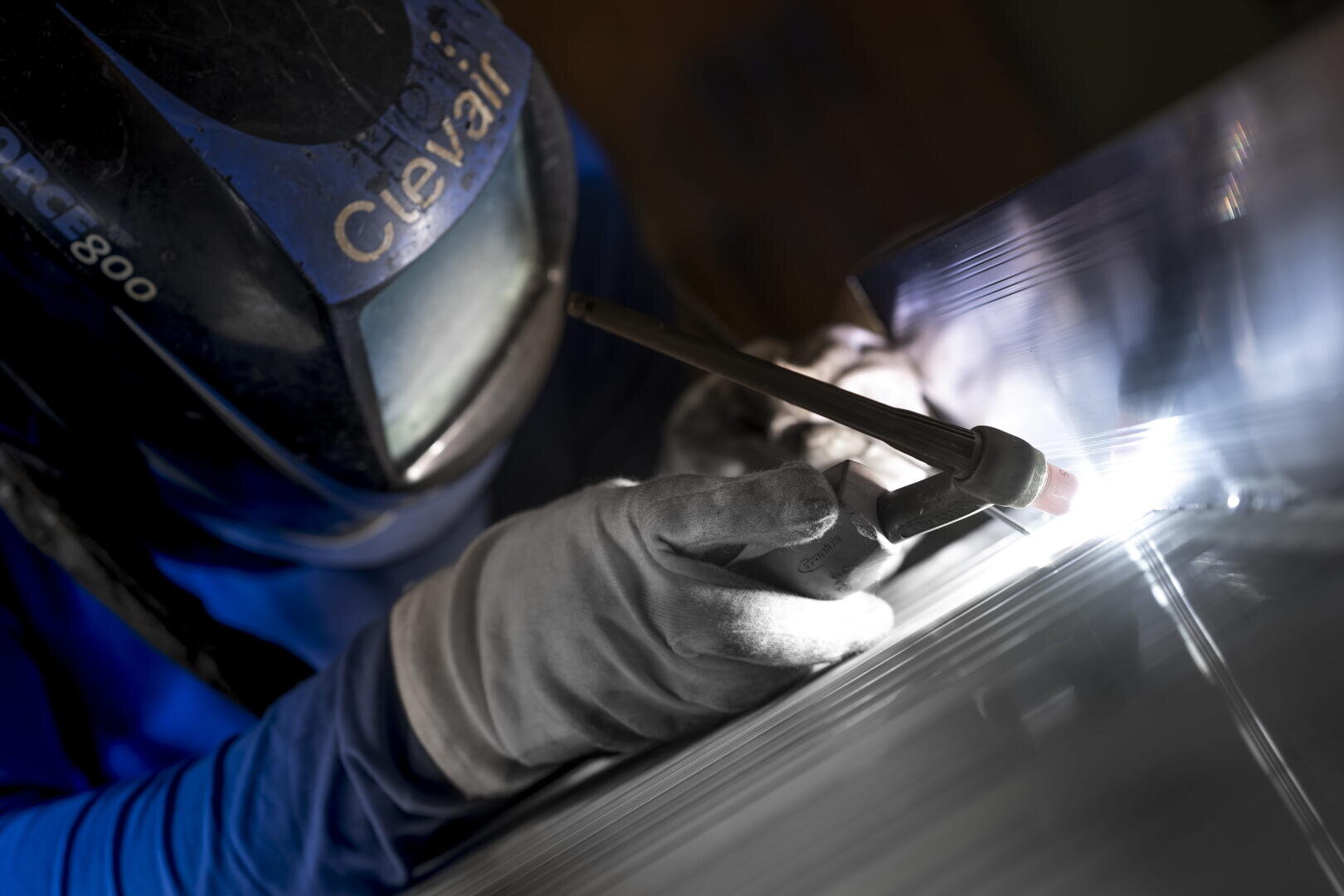 Eine Person, die einen Schweißhelm und Schutzhandschuhe trägt, bearbeitet eine Metalloberfläche mit einem WIG-Schweißbrenner.