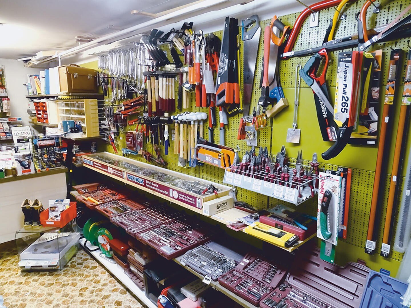 Eine Wand in einer Eisenwarenhandlung mit verschiedenen Handwerkzeugen, darunter Sägen, Schraubenschlüssel, Zangen und Schraubendreher, die ordentlich auf Steckbrettern und Regalen angeordnet sind. Werkzeugsätze und -kisten sind in den unteren Regalen ausgestellt.