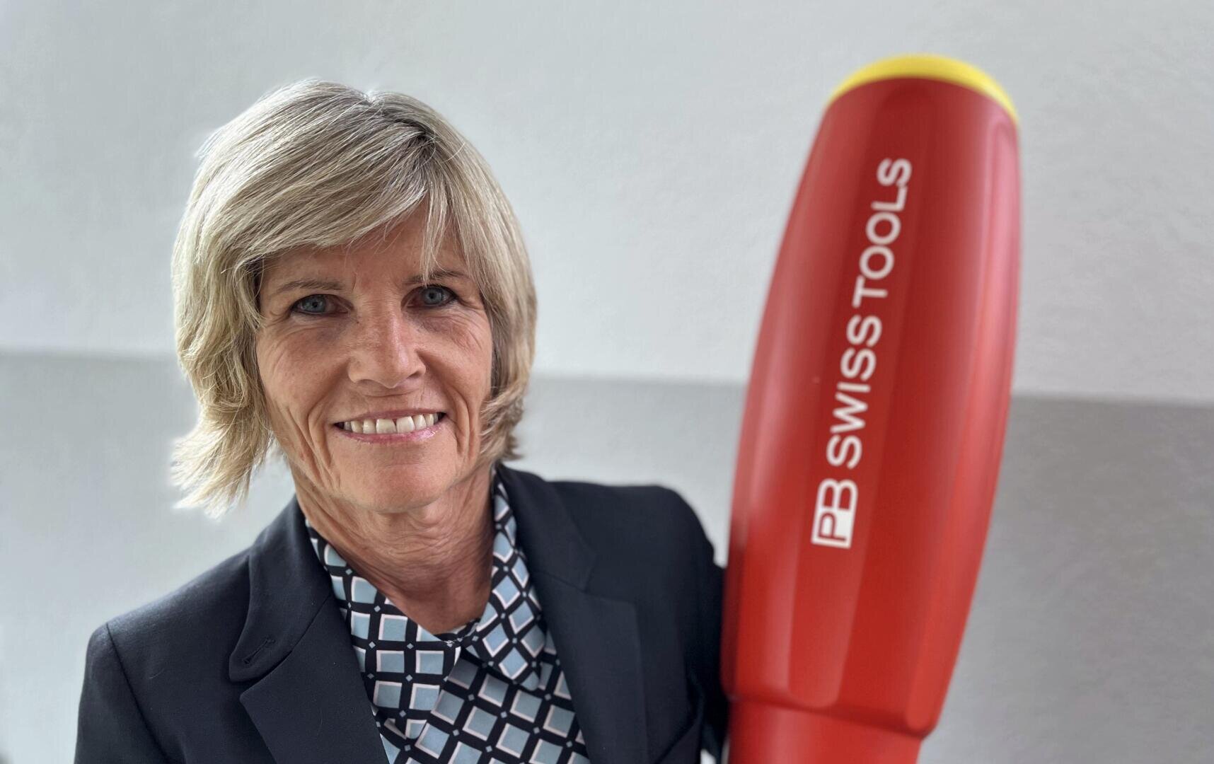 Eine lächelnde Frau in einem Blazer hält einen großen rot-gelben Gegenstand in Form eines Schraubenziehers mit der Aufschrift PB SWISS TOOLS vor einem schlichten hellen Hintergrund.