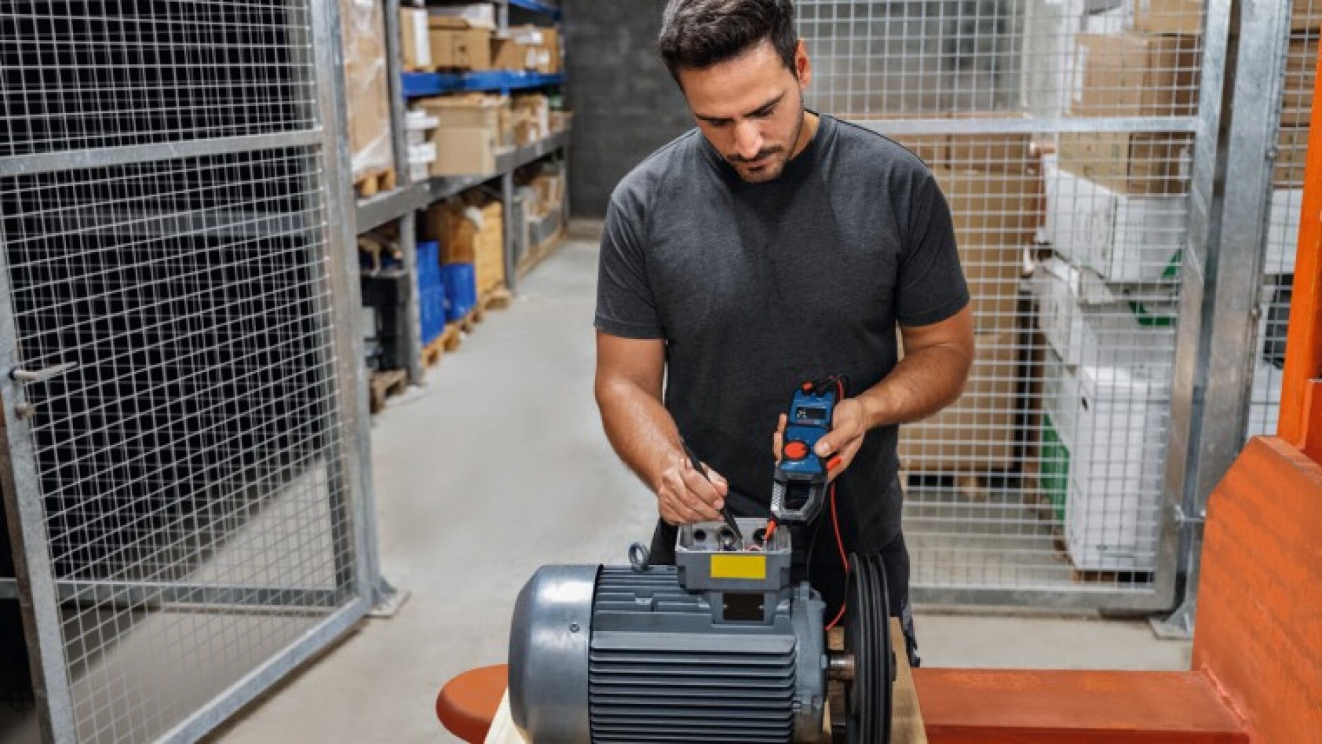 Ein Mann in einer Lagerhalle testet oder repariert mit einem Multimeter einen industriellen Elektromotor. Im Hintergrund sind Regale mit Kisten und Metallzäune zu sehen.