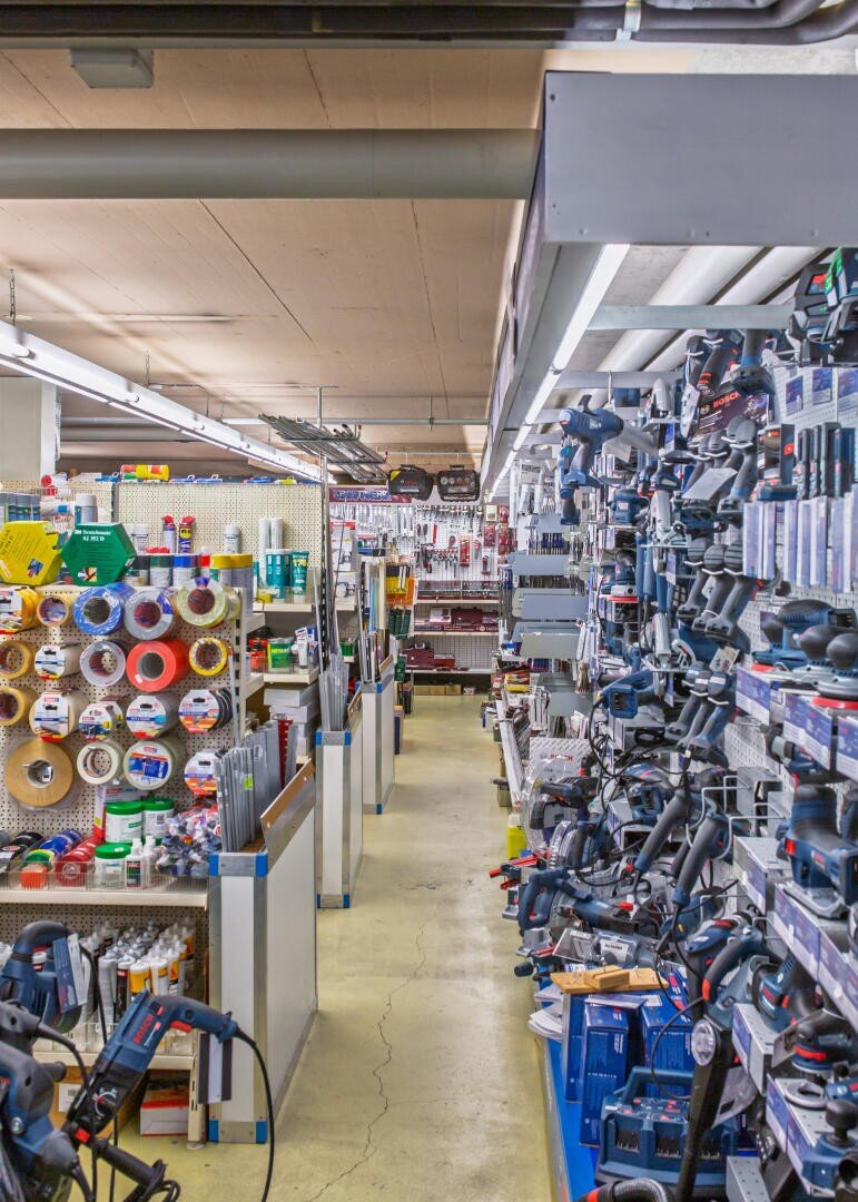 Rayon d'une quincaillerie. À droite, des étagères avec des outils électriques, à gauche, des rouleaux de ruban adhésif et d'autres articles. Le rayon est lumineux et bien approvisionné, avec plusieurs quincailleries visibles en arrière-plan.