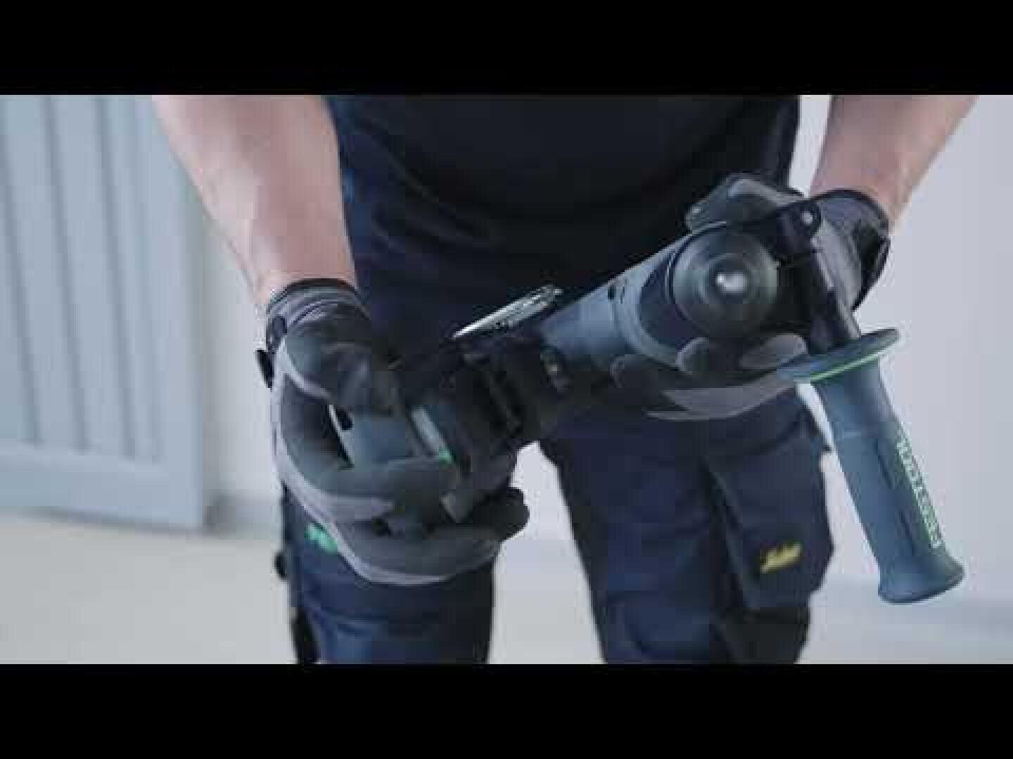 Eine Person, die Handschuhe und dunkle Arbeitskleidung trägt, hält eine Bohrmaschine mit beiden Händen fest und bereitet sich auf eine Aufgabe vor. Im Hintergrund sind eine helle Wand und eine Tür zu sehen.