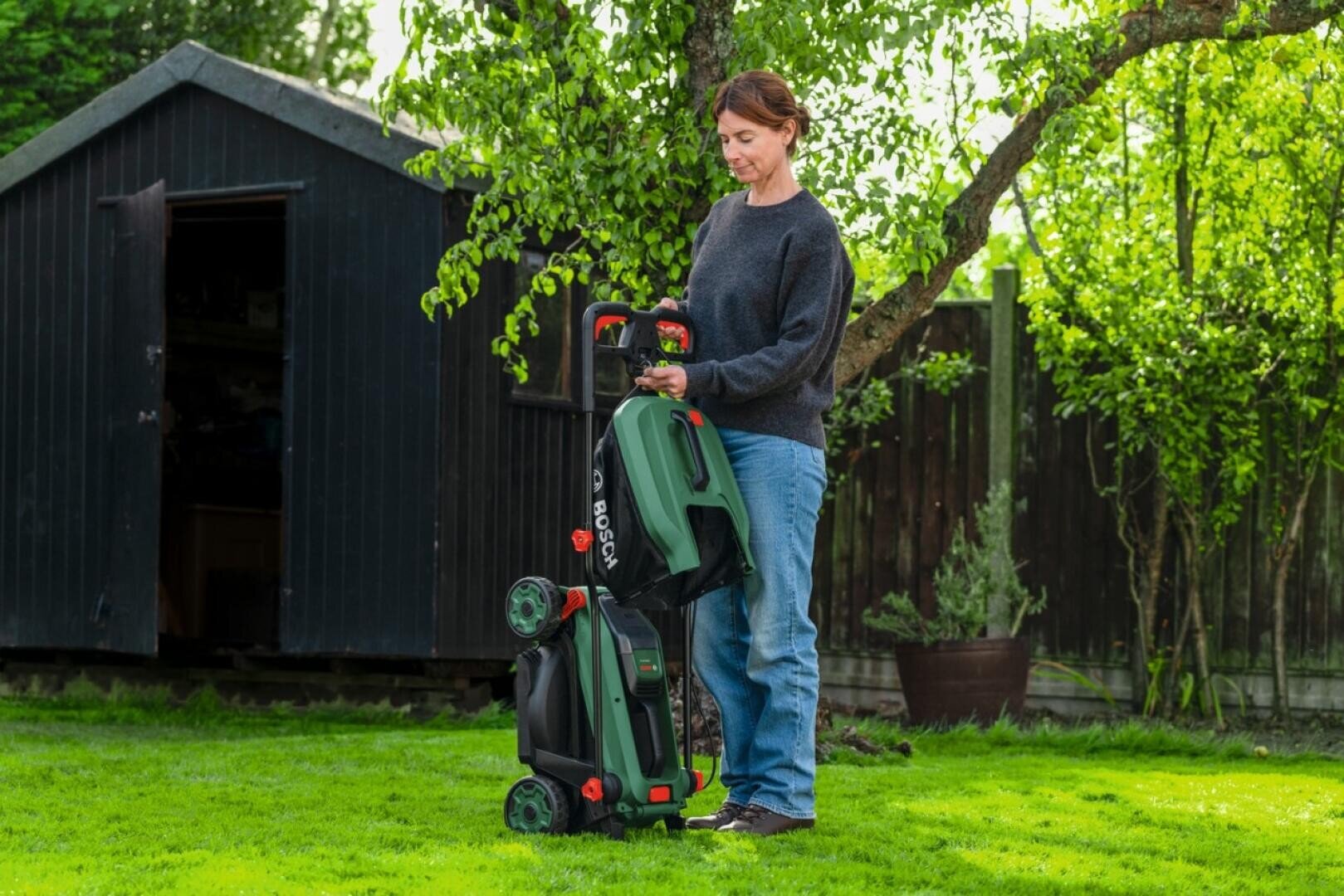 Eine Person steht auf grünem Gras und klappt einen grün-schwarzen Bosch-Rasenmäher in einem sonnigen Hinterhof neben einem Holzschuppen und einem Baum zusammen.
