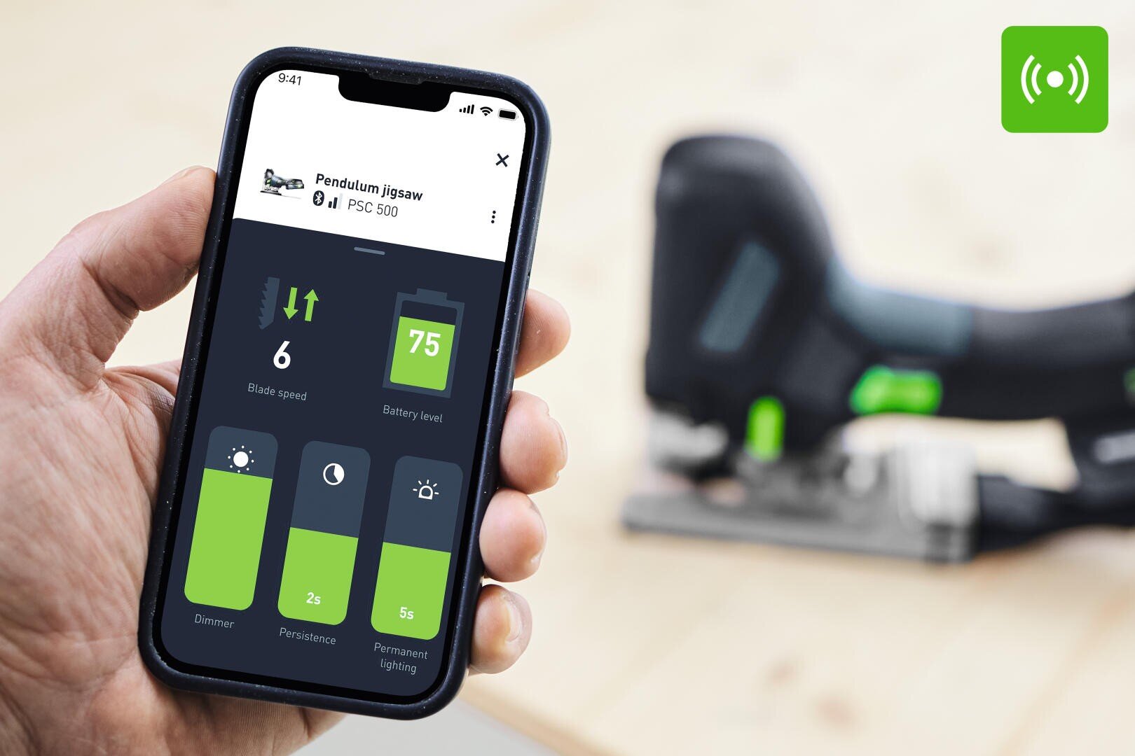 Eine Hand hält ein Smartphone, auf dem eine App zur Steuerung eines Puzzles zu sehen ist, während das Elektrowerkzeug im Hintergrund unscharf erscheint.