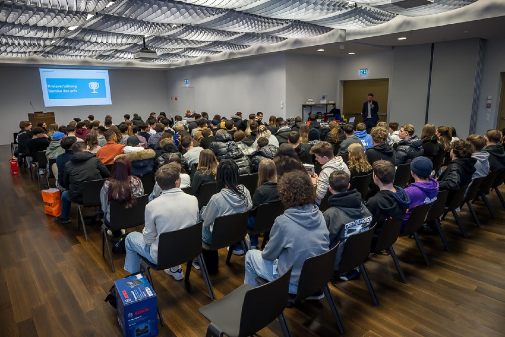 Eine große Gruppe von Studierenden sitzt in einem Konferenzraum vor einer Leinwand, auf der Preisverleihung – Remise des prix zu lesen ist.