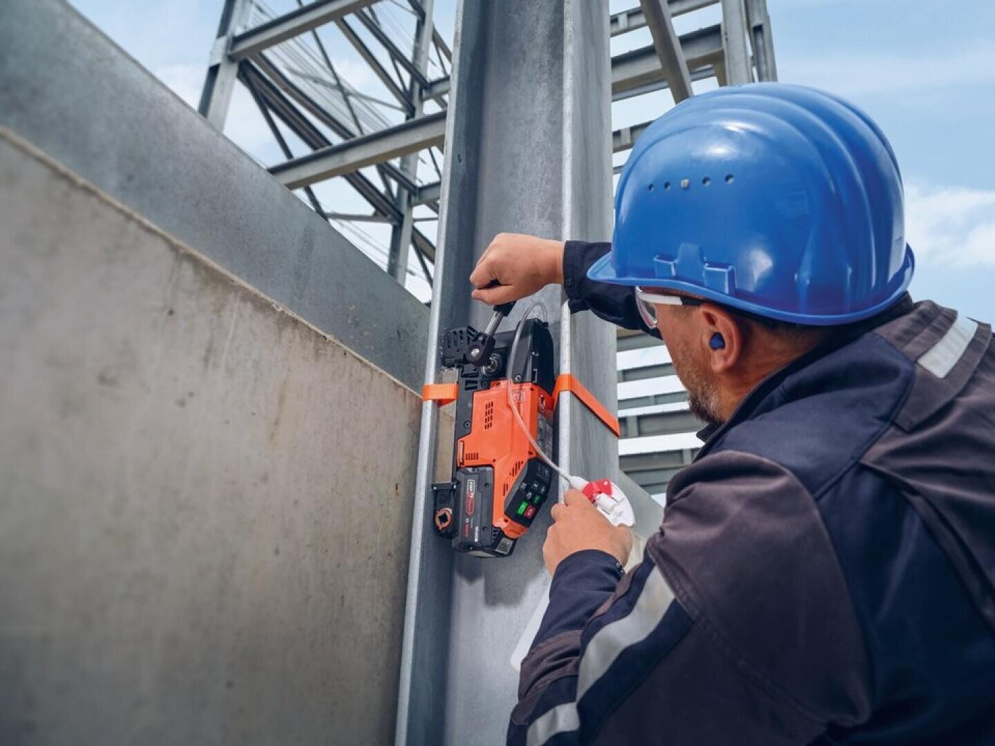 Ein Arbeiter mit einem blauen Schutzhelm befestigt auf einer Baustelle mit einem orangefarbenen Werkzeug ein Metallband um einen Stahlträger.