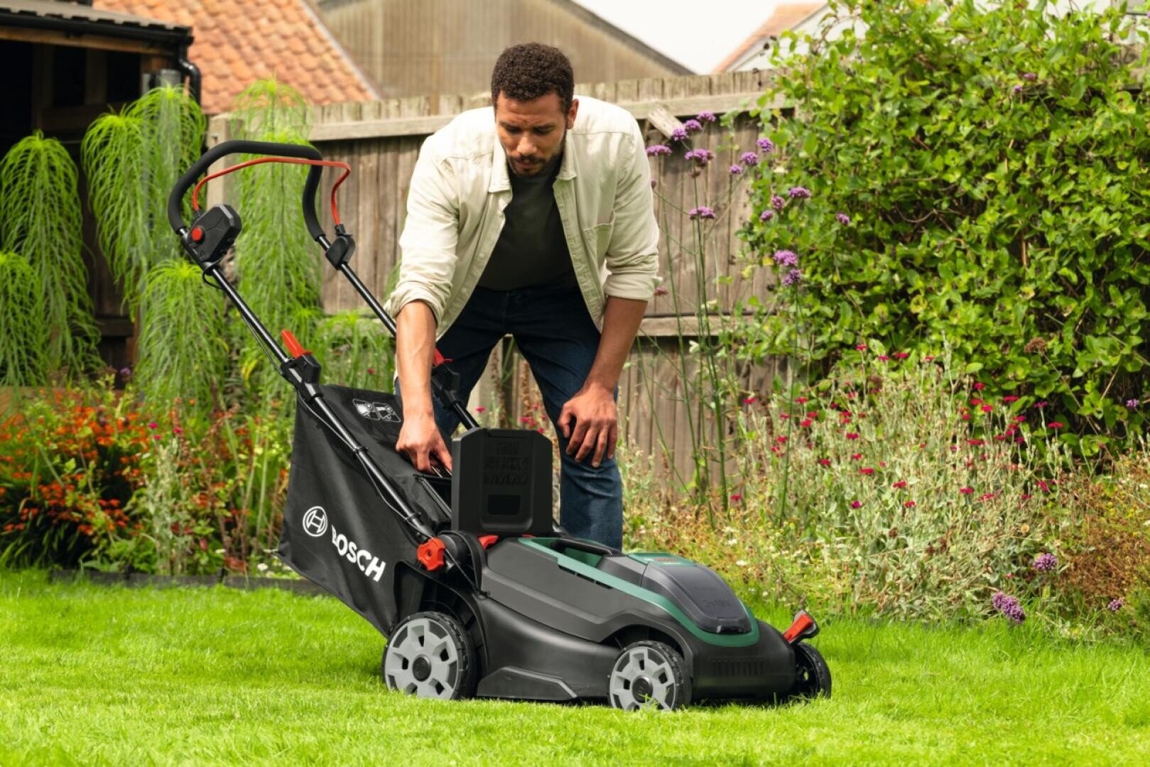 Ein Mann in einem Garten bückt sich, um einen schwarzen Bosch-Rasenmäher auf einem grünen Rasen einzustellen; im Hintergrund sind Blumen und Sträucher zu sehen, daneben steht ein Holzzaun.