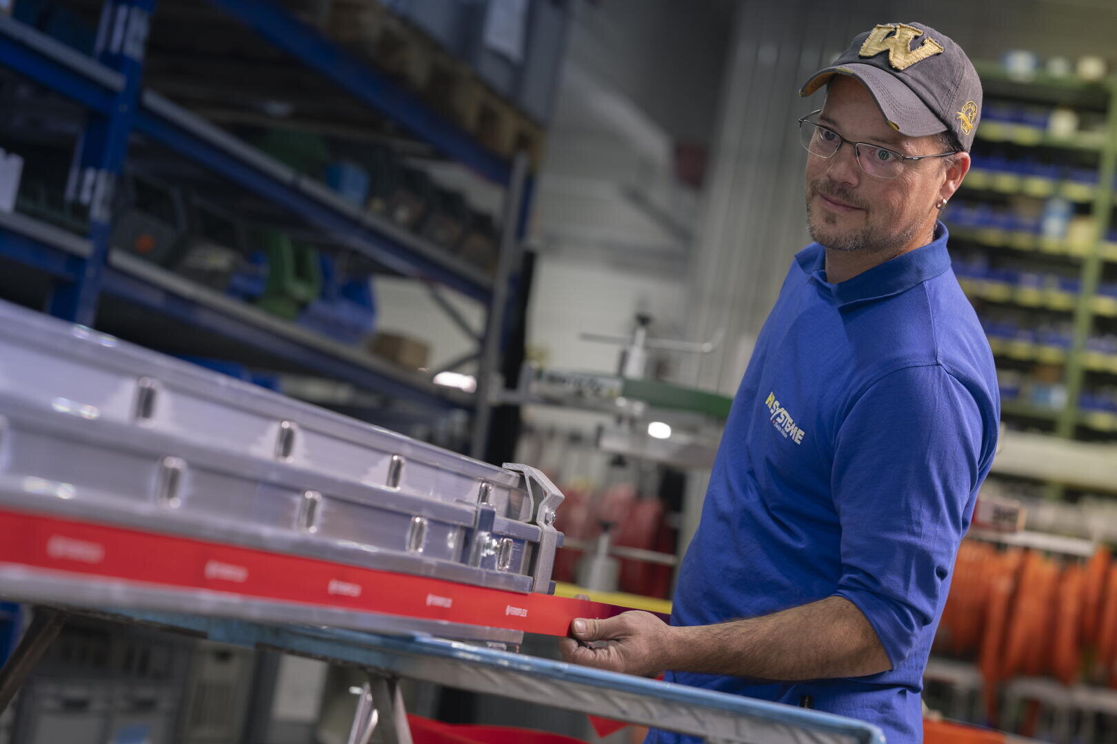 Ein Mann, der ein blaues Poloshirt und eine Mütze trägt, arbeitet an einer Werkbank in einem Lagerraum an einem Metallteil.