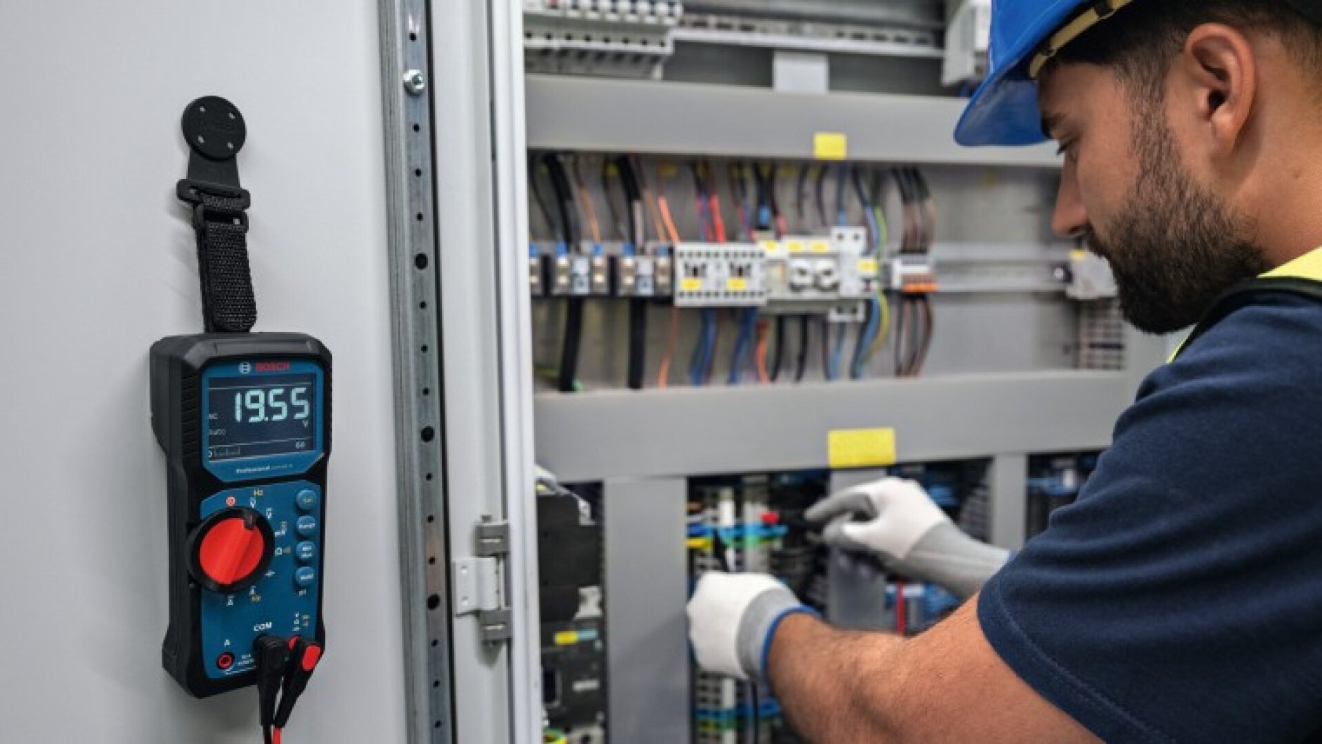Ein Elektriker mit Handschuhen und Helm arbeitet an einem Bedienfeld mit verschiedenen Kabeln und Komponenten. An der Schalttafeltür ist ein Digitalmultimeter mit der Anzeige „19,55“ angebracht.