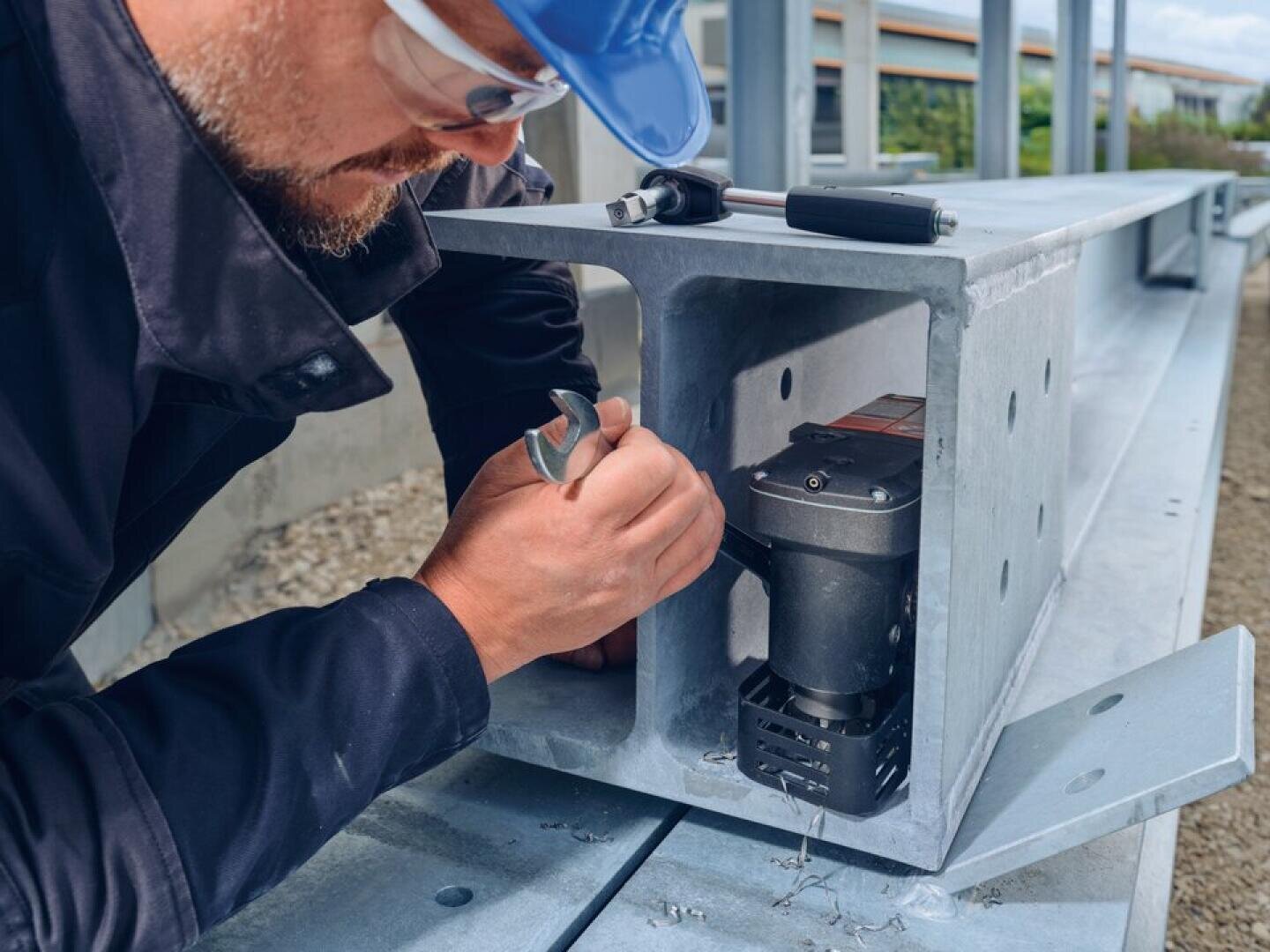 Ein Arbeiter, der eine Schutzbrille und einen blauen Schutzhelm trägt, zieht mit einem Schraubenschlüssel eine Schraube in einer großen Metallkonstruktion im Freien fest. Im Hintergrund sind Werkzeuge und Metallträger zu sehen.