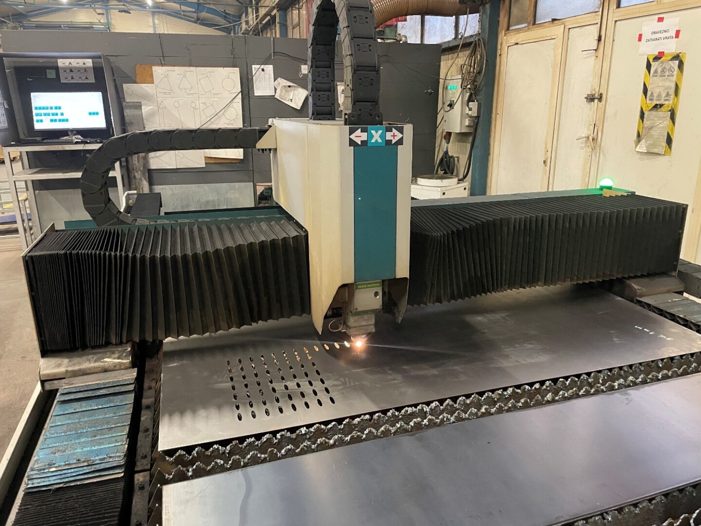 Eine CNC-Laserschneidmaschine schneidet in einer Industriewerkstatt präzise Löcher in ein großes Metallblech. Im Hintergrund sind Bedienfelder und Zeichnungen zu sehen.