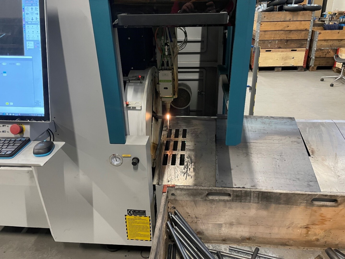 Eine CNC-Maschine schneidet mit einem Laser präzise Schlitze in ein Blech. In der Nähe stapeln sich Metallstangen und Holzkisten. Auf der linken Seite der Maschine sind ein Computerbildschirm und Bedienelemente zu sehen.