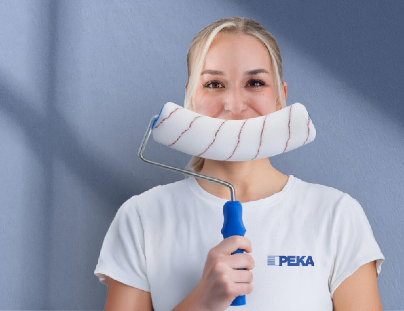 Eine Frau in einem weißen PEKA-Shirt hält eine Farbrolle waagerecht unter ihre Nase, so dass es wie ein breites Lächeln aussieht, während sie vor einer blauen Wand steht.