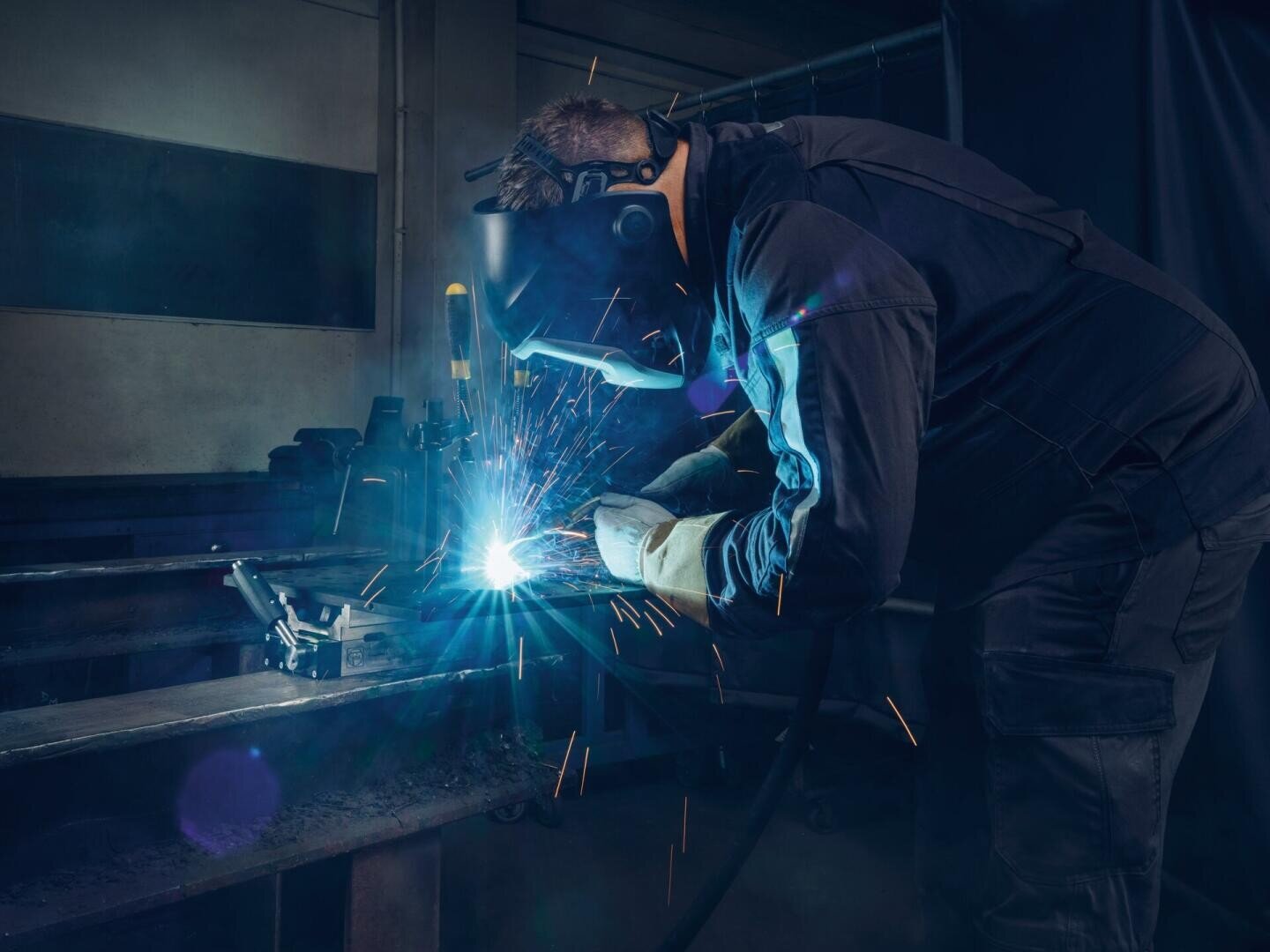 Eine Person, die Schutzkleidung und eine Schweißermaske trägt, schweißt in einer Industriehalle Metall, wobei hellblaue Funken sprühen.
