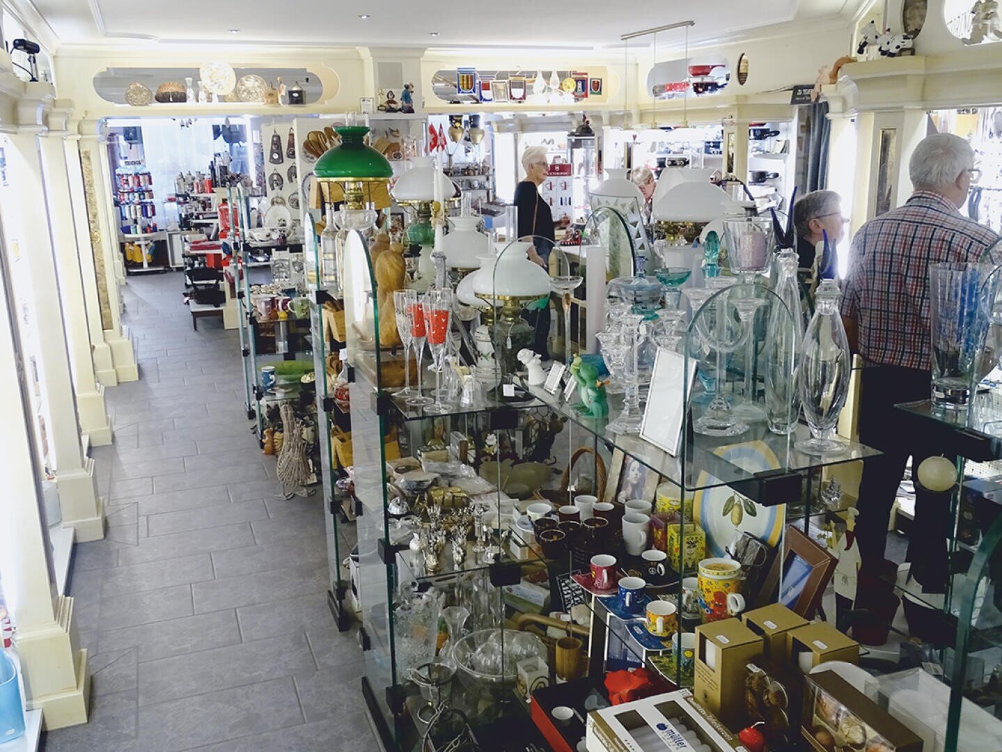 Ein hell erleuchtetes Ladeninnere mit Regalen und Auslagen, die mit Glaswaren, Keramik, Lampen und verschiedenen Dekorationsartikeln gefüllt sind. Mehrere Kunden stöbern in den Artikeln.