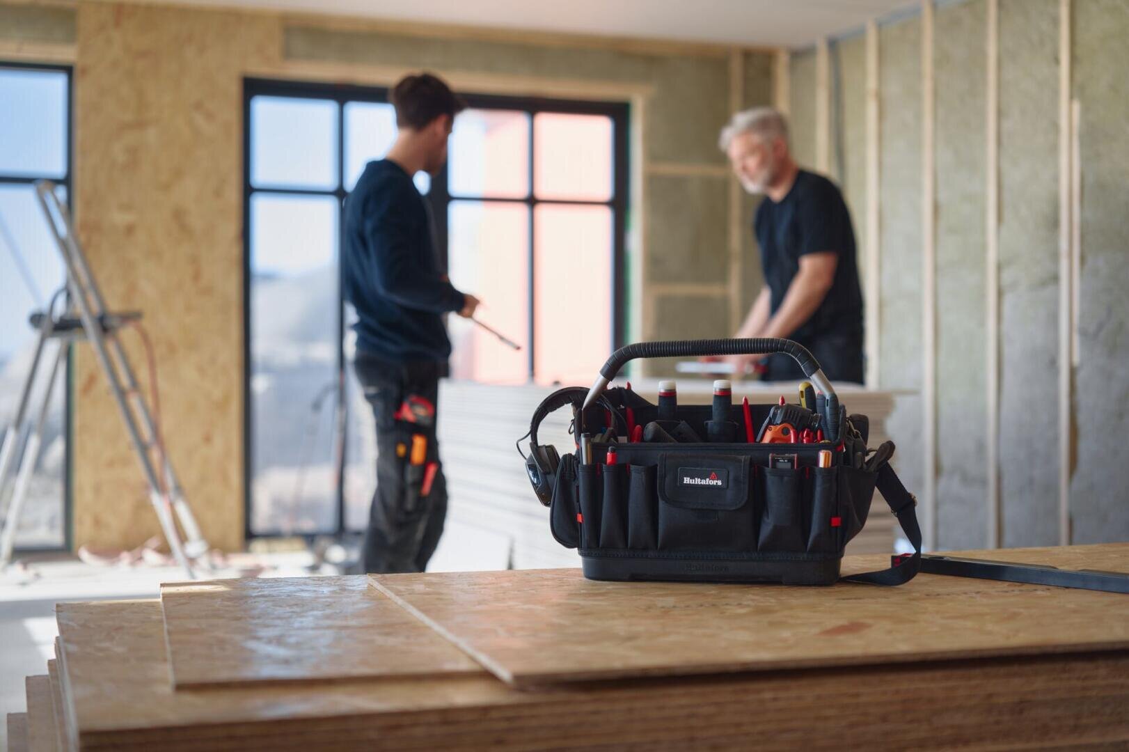 In einem im Bau befindlichen Raum steht eine Werkzeugtasche auf Sperrholzplatten, während im Hintergrund in der Nähe großer Fenster zwei verschwommene Arbeiter über Baumaterialien diskutieren.