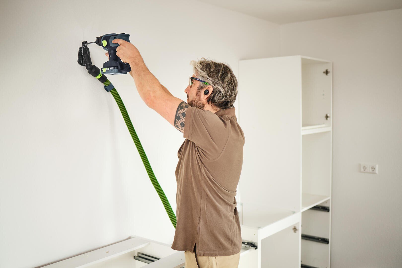 Eine Person, die eine Schutzbrille und einen Gehörschutz trägt, benutzt eine Bohrmaschine mit einem daran befestigten grünen Schlauch, um in eine weiße Wand in einem Raum zu bohren, in dem teilweise montierte weiße Schränke stehen.