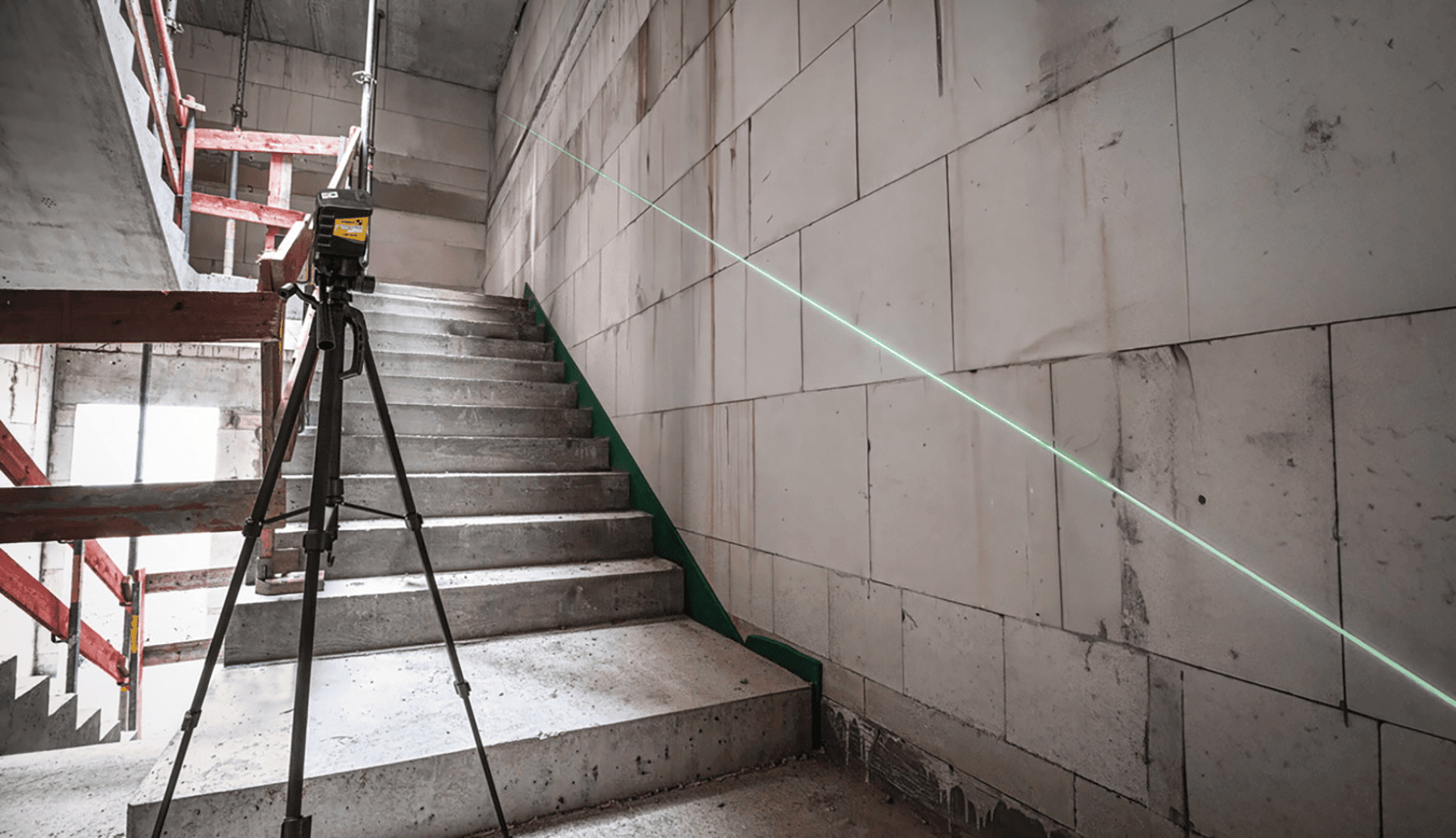 Ein Laser-Nivelliergerät auf einem Stativ projiziert eine grüne diagonale Linie auf eine Betonblockwand neben einer im Bau befindlichen Treppe.