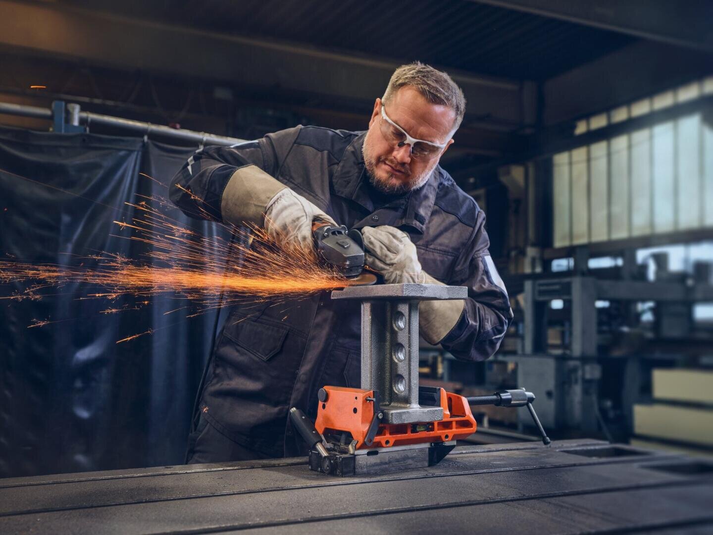 Ein Mann, der eine Schutzbrille und Handschuhe trägt, schneidet mit einem Winkelschleifer, der Funken sprüht, ein Metallstück, das auf einer Werkbank in einer Industriewerkstatt befestigt ist.