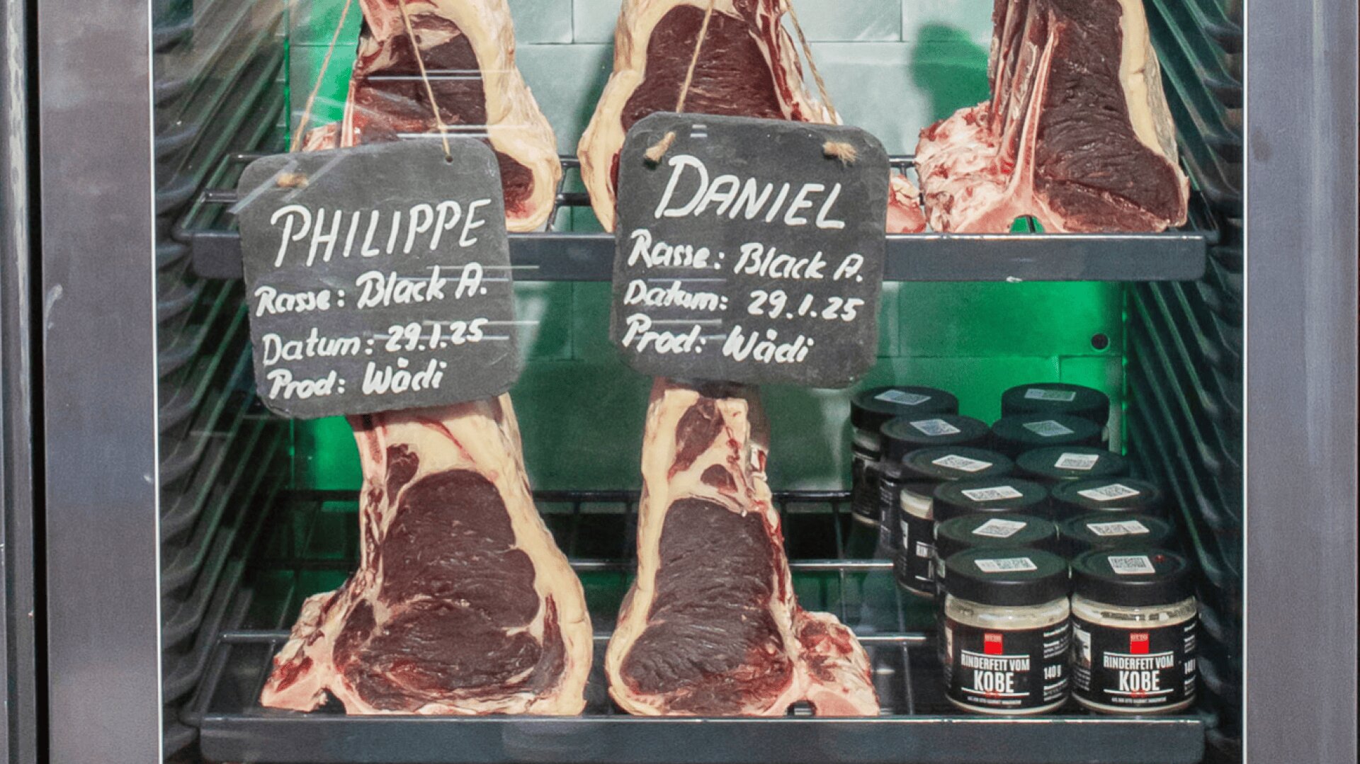 Große Stücke marmorierten Rindfleischs mit den Aufschriften „Philippe“ und „Daniel“ liegen in den Regalen einer Fleischkühltruhe, neben Gewürzgläsern mit der Aufschrift „Kobe“. Schwarze Schilder geben Auskunft über Herkunft und Datum des Fleisches.