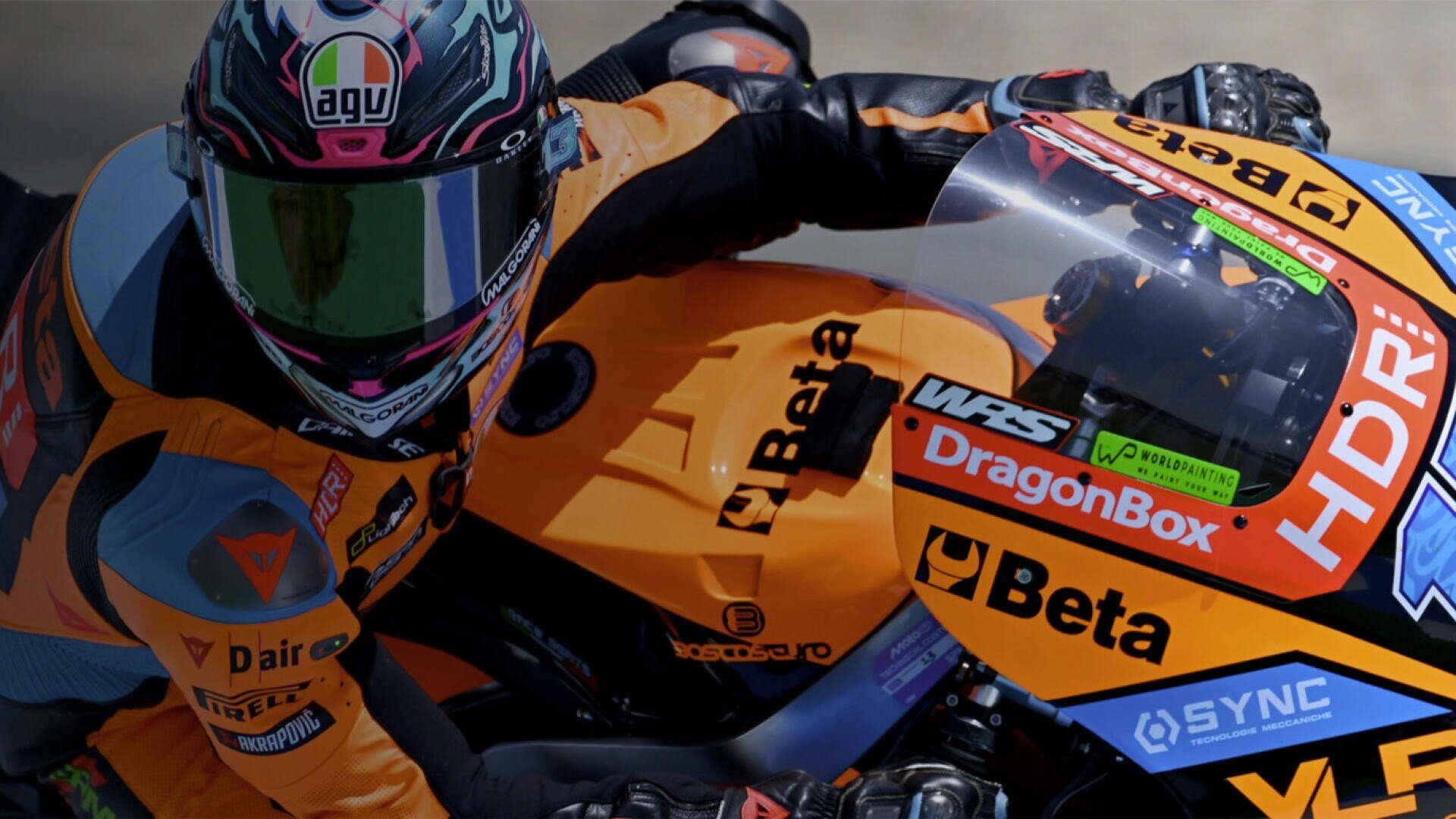 Ein Motorradrennfahrer in orangefarbener Rennkleidung und mit orangefarbenem Helm fährt auf einem orangefarbenen Motorrad, das mit Sponsorenlogos übersät ist, und neigt sich während des Rennens stark zur Seite.