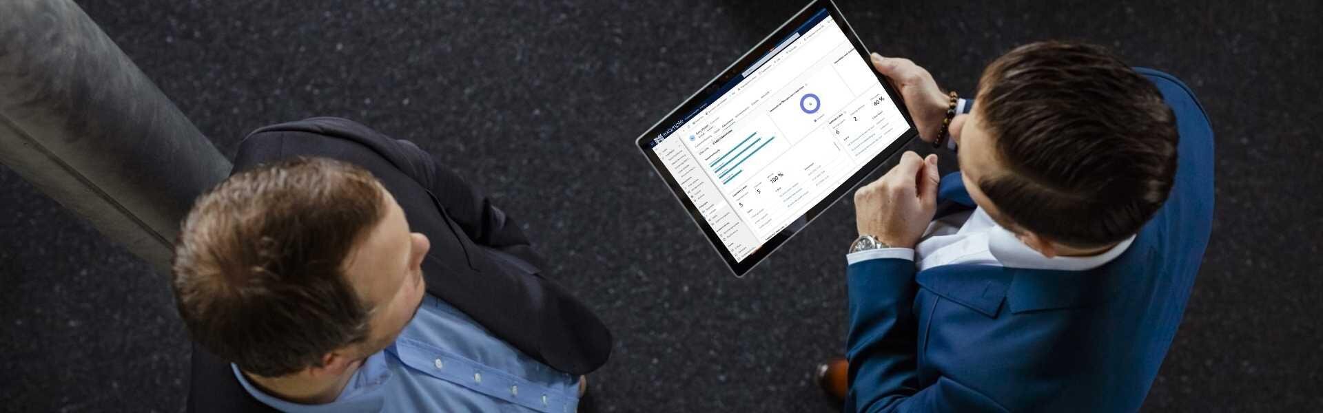 Zwei Männer in Geschäftskleidung stehen zusammen und blicken auf ein Tablet, auf dem Grafiken und Analysedaten angezeigt werden, die von oben auf einem dunklen, strukturierten Boden zu sehen sind.