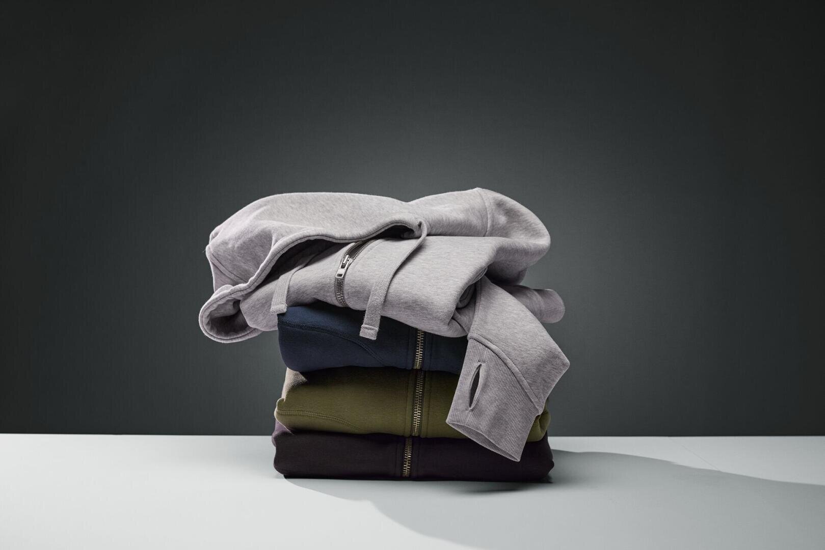 Ein ordentlich gefalteter Stapel aus drei Hoodies in Grau, Dunkelblau und Olivgrün liegt auf einer weißen Fläche vor einem dunklen Hintergrund.