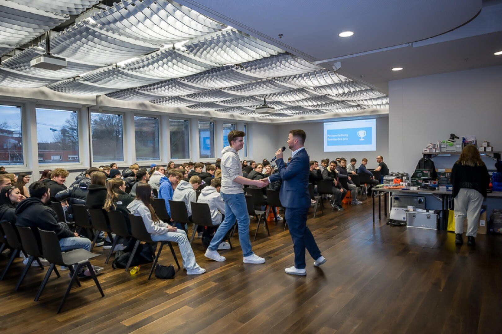 Ein Mann im Anzug übergibt einem jungen Mann in Jeans vor einem Publikum in einem modernen Konferenzraum ein Mikrofon; auf einer Leinwand hinter ihnen wird eine Präsentation angezeigt.