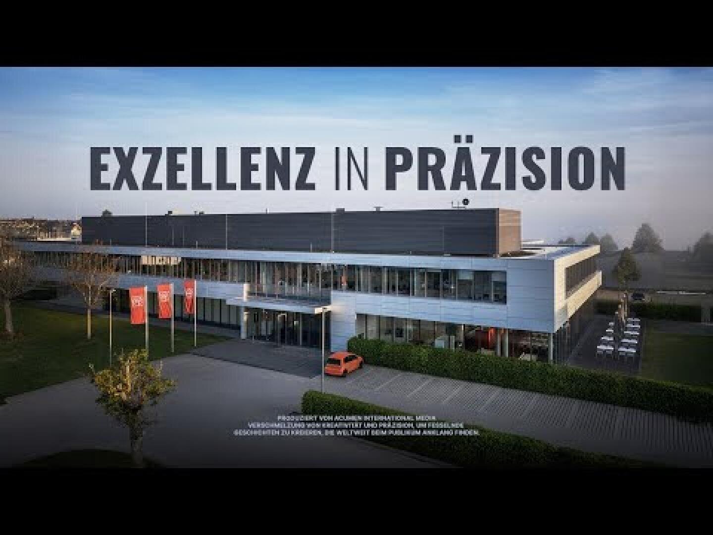 Ein modernes, graues Gebäude mit großen Fenstern, umgeben von Grünflächen und geparkten Autos. Darüber ist in fetter Schrift „EXZELLENZ IN PRÄZISION“ zu lesen. Die Szene wirkt geschäftsmäßig und professionell.