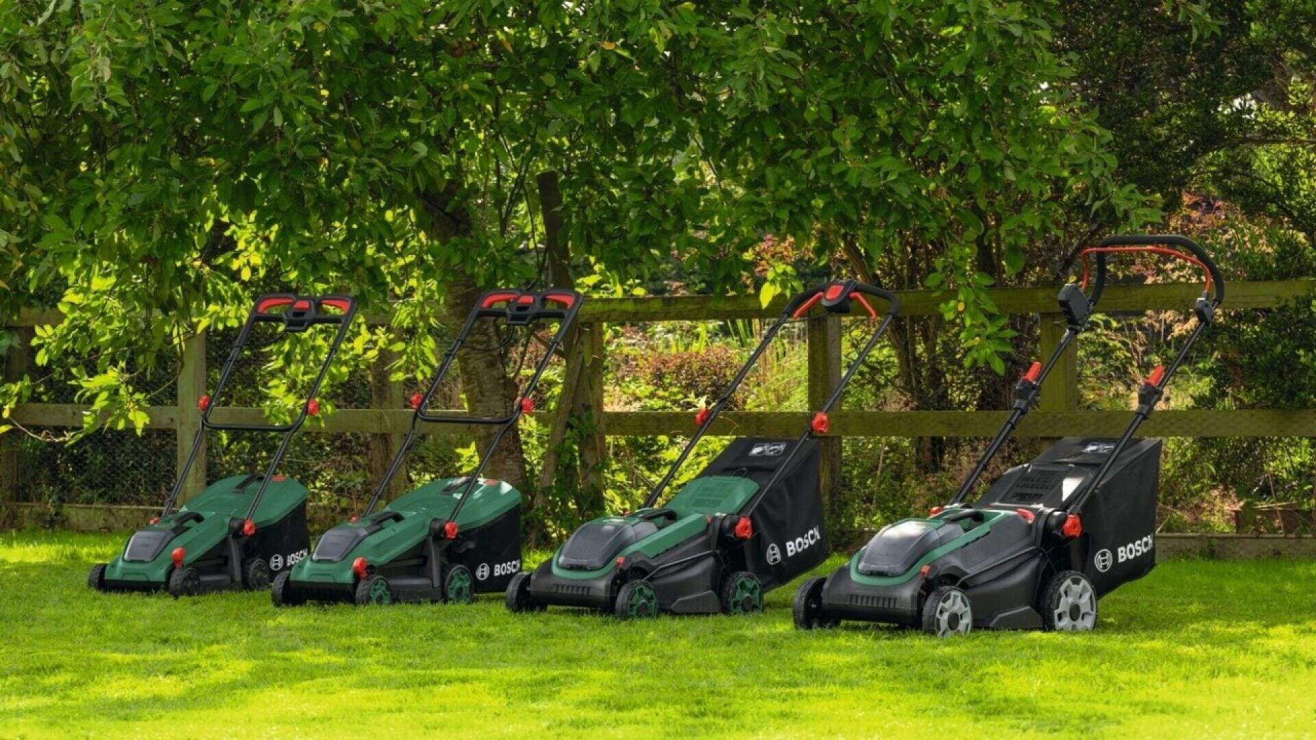 Vier grüne Bosch-Rasenmäher unterschiedlicher Größe stehen auf einer Rasenfläche in der Nähe eines Holzzauns, im Hintergrund sind Bäume und Sträucher zu sehen.