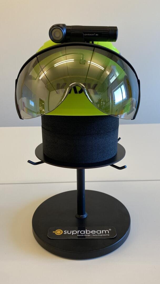 Ein leuchtend gelber Schutzhelm mit getöntem Visier und einer oben angebrachten Suprabeam-Stirnlampe, ausgestellt auf einem schwarzen Ständer.