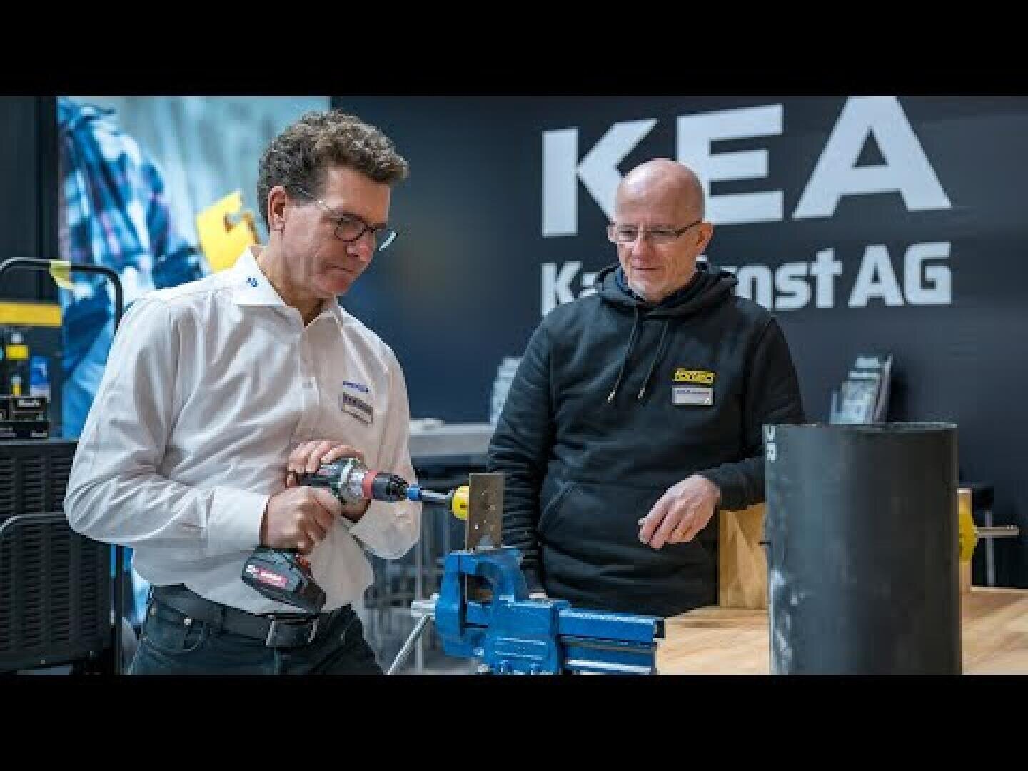 Zwei Männer stehen an einer Werkbank; einer bearbeitet mit einer Bohrmaschine ein Metallteil, das in einem blauen Schraubstock eingespannt ist. Beide tragen Namensschilder und eine Brille. Auf einer schwarzen Wand hinter ihnen sind die Buchstaben „KEA“ und weiterer, teilweise verdeckter Text zu sehen.