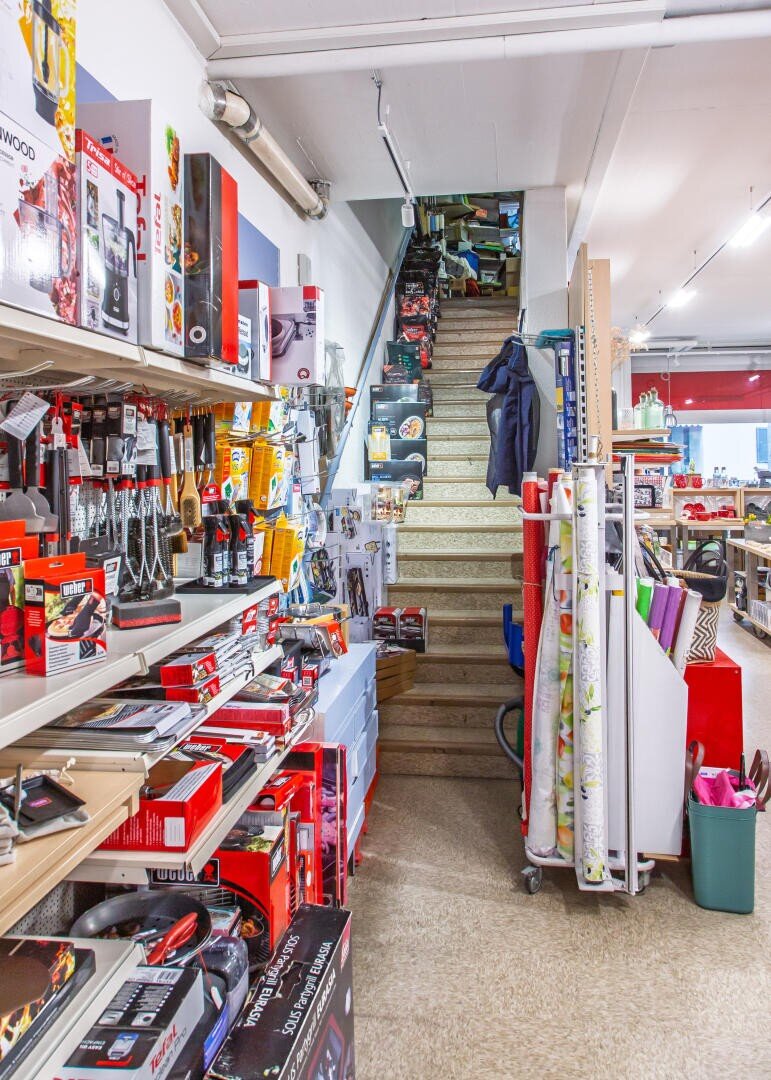 L'intérieur d'une quincaillerie avec des étagères remplies d'outils et d'appareils de cuisine (à gauche), des rouleaux de papier cadeau (à droite) et un escalier central menant à l'étage. Le magasin est lumineux et bien organisé.