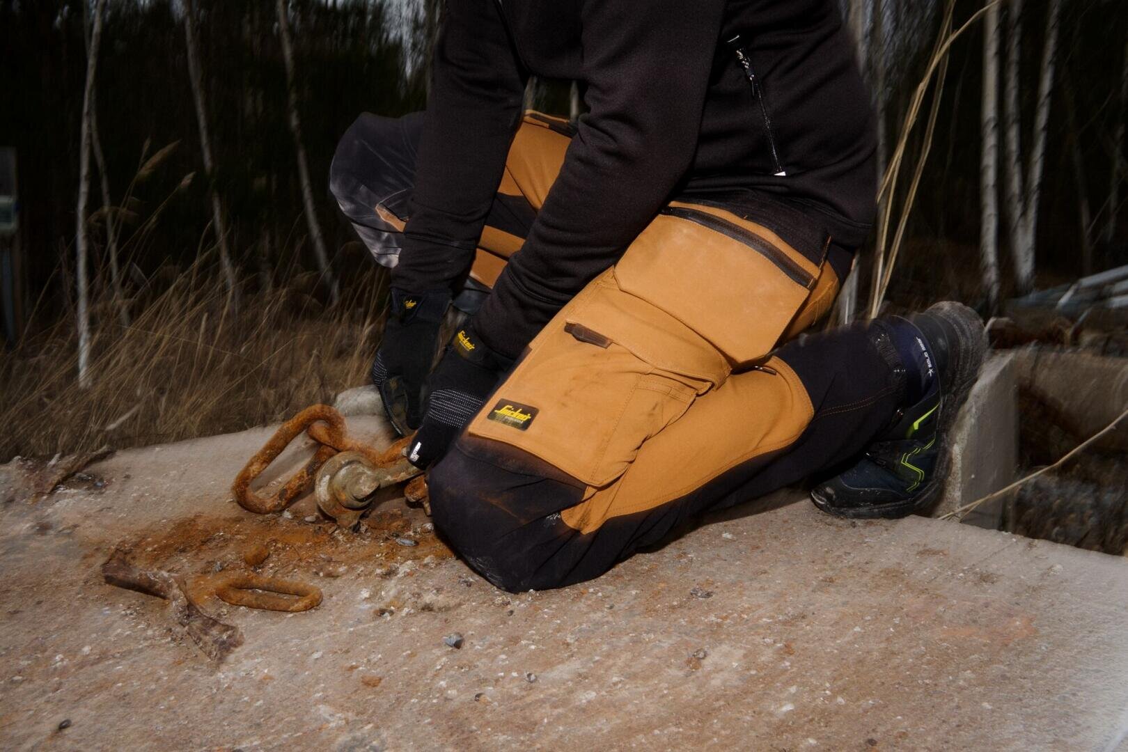 Eine Person in braunen Arbeitshosen und schwarzen Handschuhen kniet draußen auf einer Betonfläche und bearbeitet mit Werkzeugen rostige Metallschlaufen.