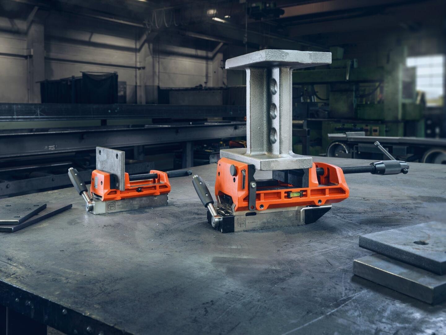 Zwei orange-schwarze magnetische Schweißklemmen halten Metallteile auf einer Werkbank in einer Industriewerkstatt fest, im Hintergrund sind Maschinen und Geräte zu sehen.
