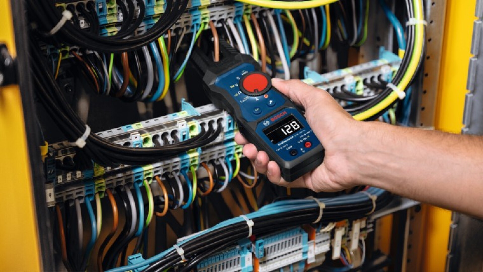Eine Person hält ein digitales Zangenmessgerät in der Hand und misst damit den elektrischen Strom in einem Bedienfeld mit zahlreichen Drähten und Kabeln in verschiedenen Farben. Das Display zeigt einen Wert von 128 an.
