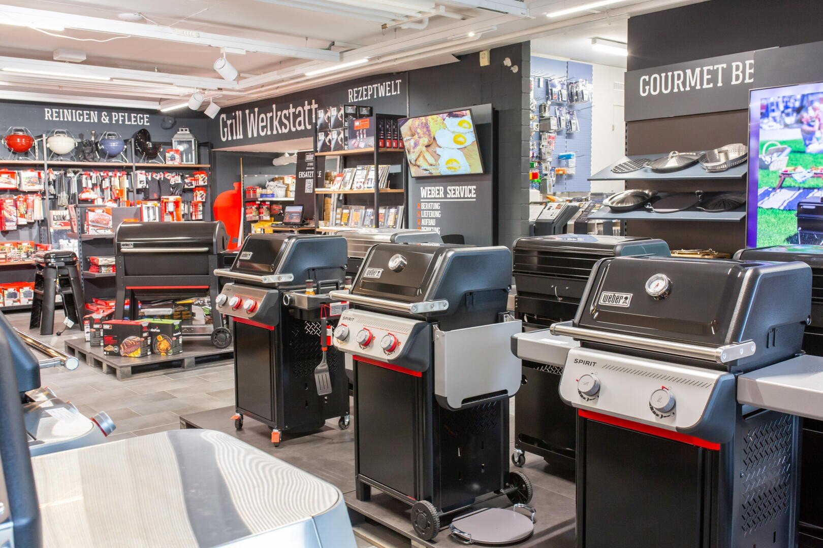Im Ladeninneren sind mehrere schwarze Gasgrills aufgereiht, umgeben von Grillzubehör, Grillwerkzeugen und Werbedisplays. Die Regale sind mit Produkten gefüllt, und an den Wänden sind Schilder in deutscher Sprache zu sehen.