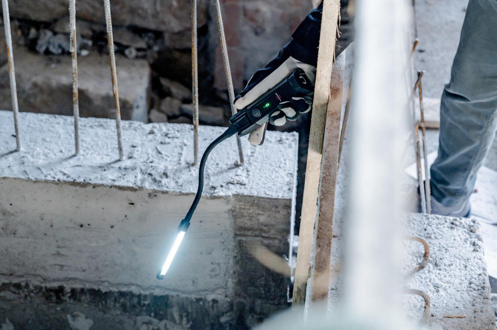 Eine mit einem Handschuh bekleidete Hand hält eine flexible Inspektionskamera mit Beleuchtung und richtet sie in ein Betonloch auf einer Baustelle, in dem Stahlbewehrungen freiliegen.