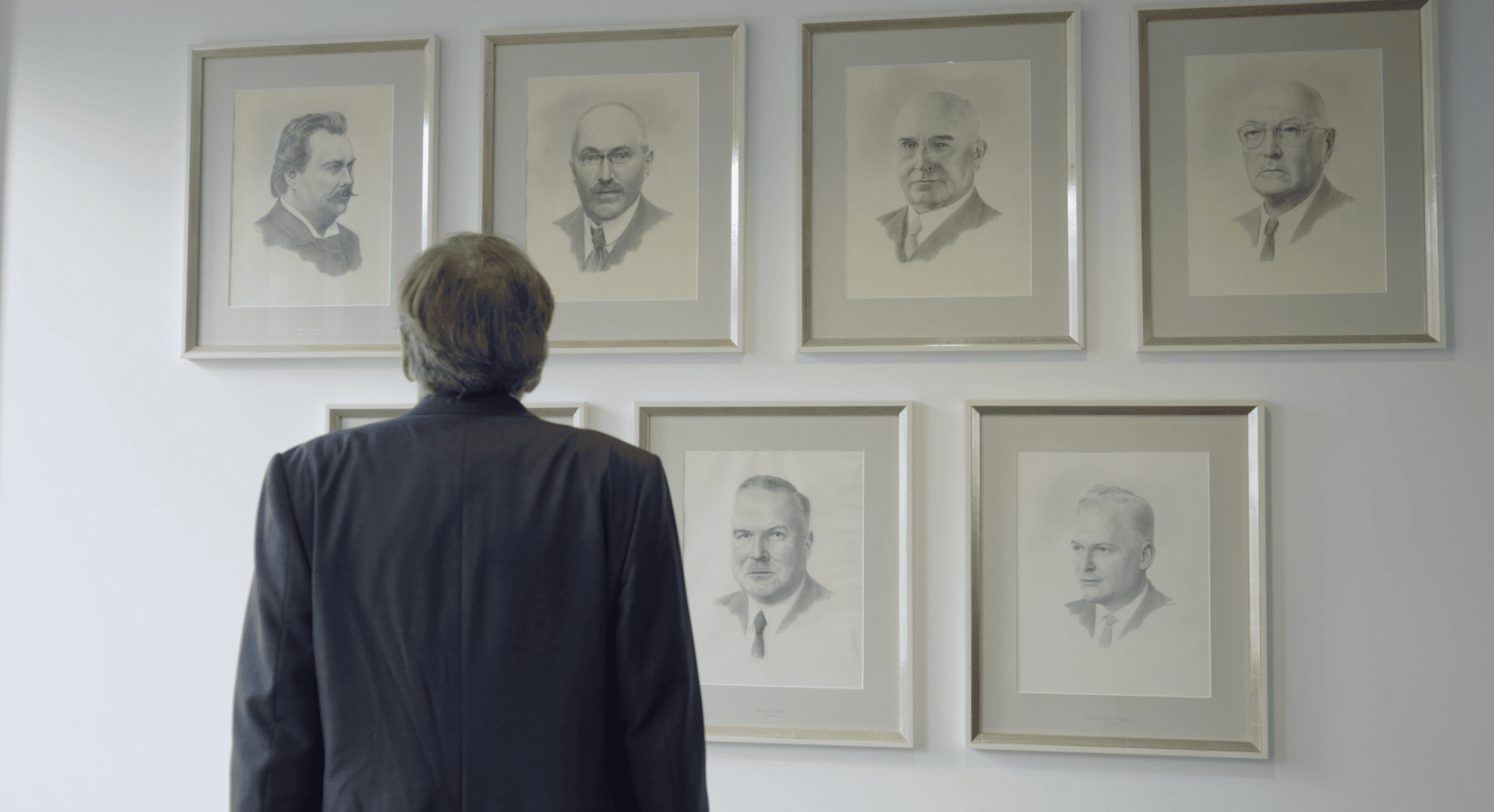 Ein Mann im Anzug steht mit dem Rücken zur Kamera und blickt auf eine Wand, an der gerahmte Schwarz-Weiß-Porträts von sechs Männern hängen.