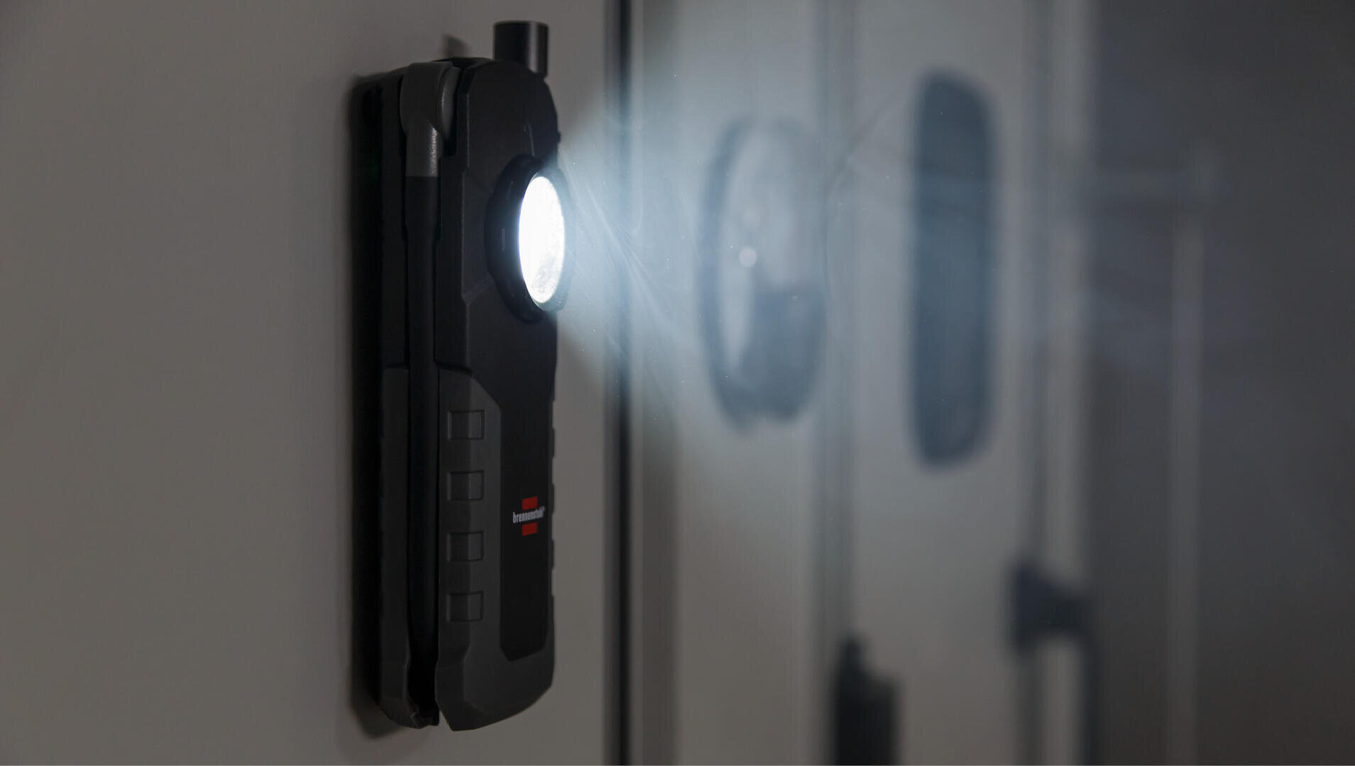 Eine schwarze Taschenlampe mit einem hellen Lichtstrahl ist magnetisch an einer grauen Metalloberfläche befestigt und beleuchtet den Bereich vor ihr.