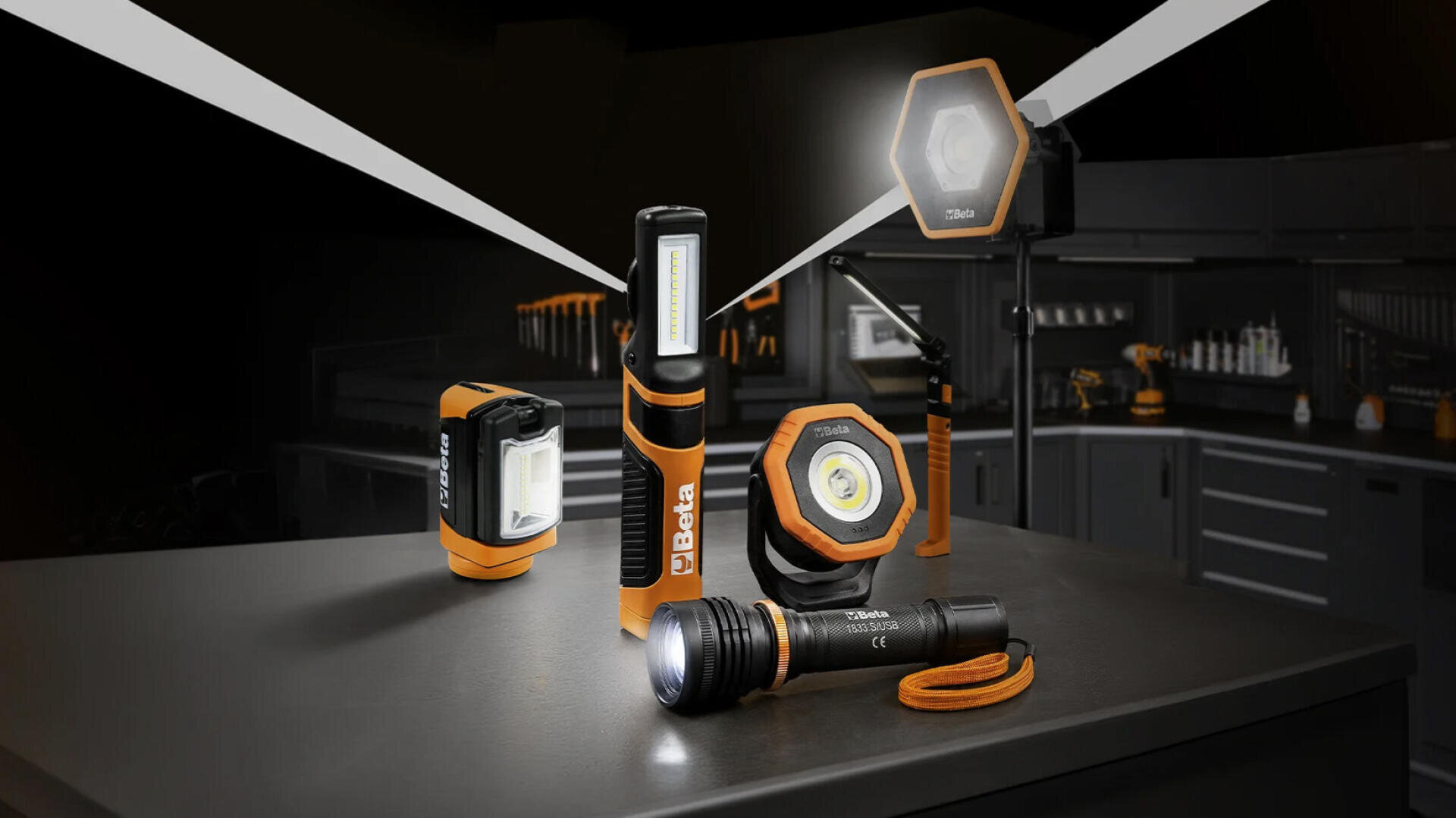 Auf einer dunklen Werkbank in einer Werkstatt sind fünf orange-schwarze LED-Arbeitsleuchten zu sehen, aus denen Lichtstrahlen scheinen. Im Hintergrund sind verschiedene Werkzeuge und Schränke zu erkennen, die eine industrielle Atmosphäre schaffen.