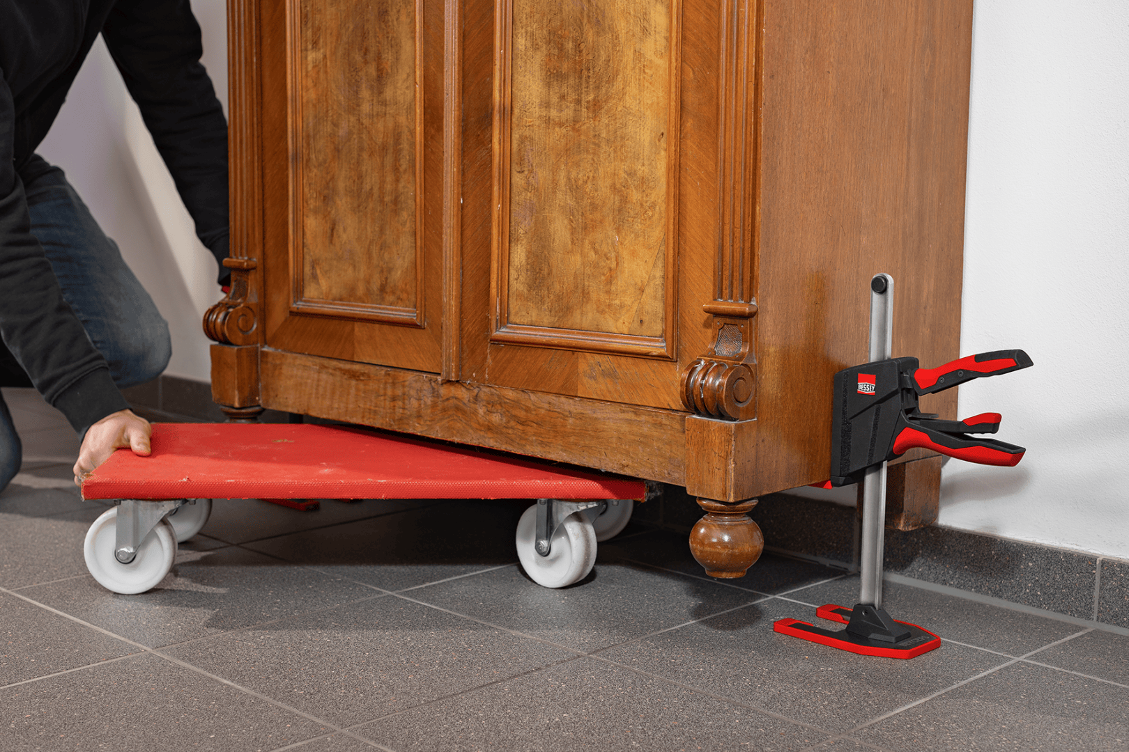 Eine Person benutzt einen roten Rollwagen und ein Hebewerkzeug mit einem Griff und einer Klemme, um einen schweren Holzschrank über einen Fliesenboden zu bewegen.