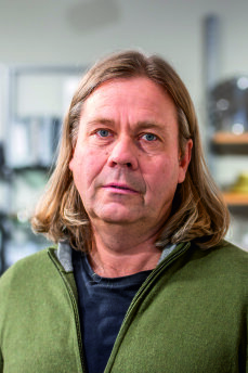 Eine Person mittleren Alters mit langem, hellbraunem Haar und blauen Augen trägt einen grünen Pullover über einem schwarzen Hemd und steht in einem Gebäude mit Regalen und verschwommenen Objekten im Hintergrund.